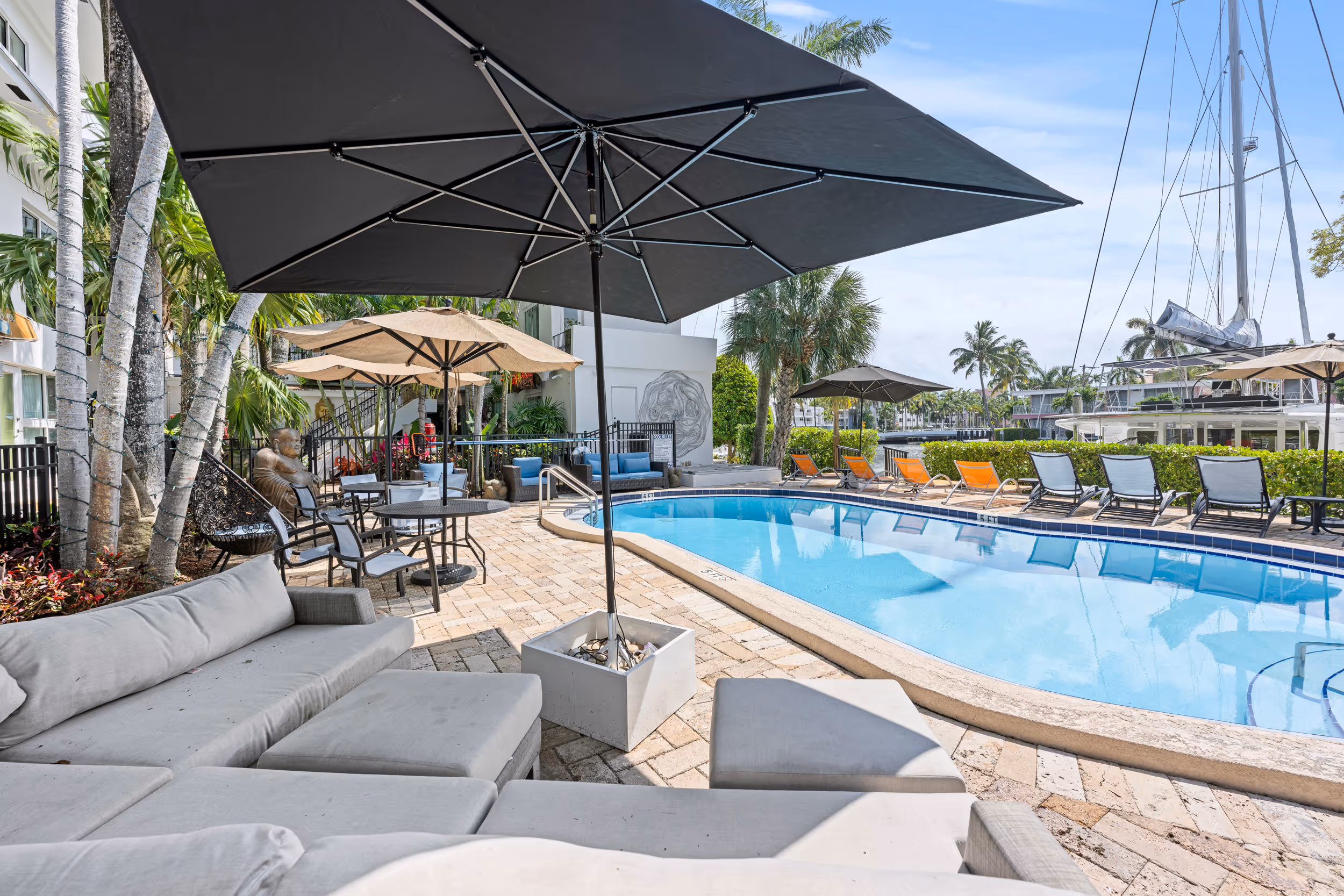 Outdoor pool area with lounge chairs, umbrellas, and seating near palm trees and a docked yacht.