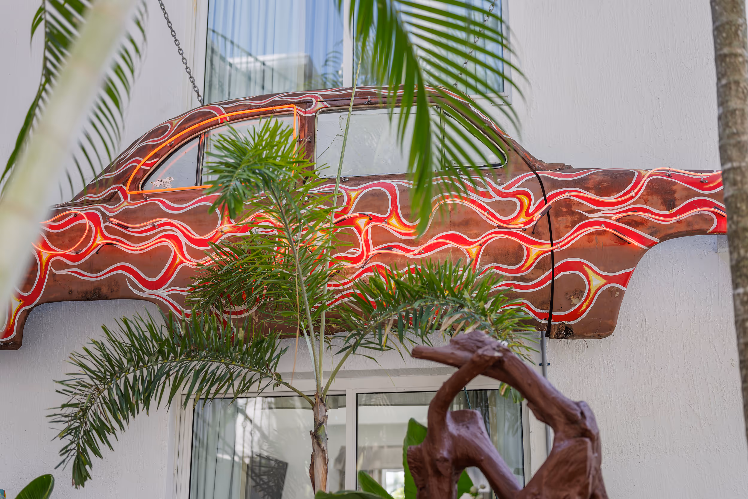 Rusty car body with red, yellow, and white flame designs mounted on a white wall behind green palm leaves.