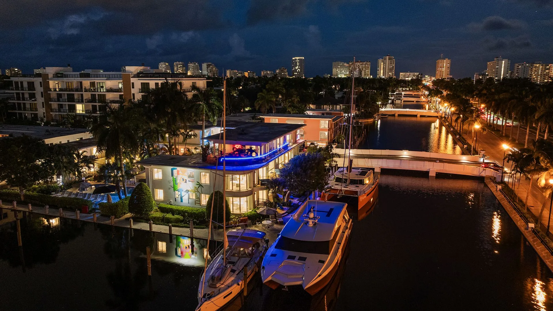 Nighttime aerial view of illuminated waterfront buildings, boats docked along a canal, and city skyline in the background.