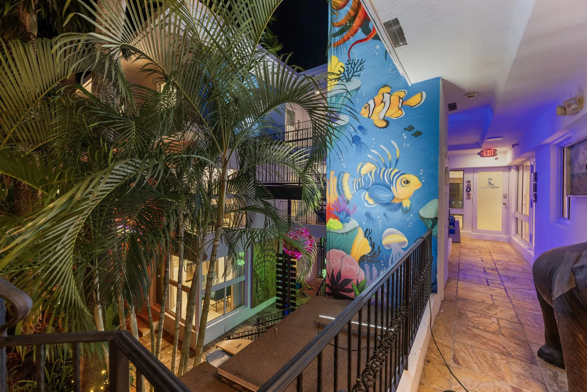 Night view of a balcony walkway with ornate railing, tropical palm trees, and a colorful underwater-themed mural featuring clownfish and coral.
