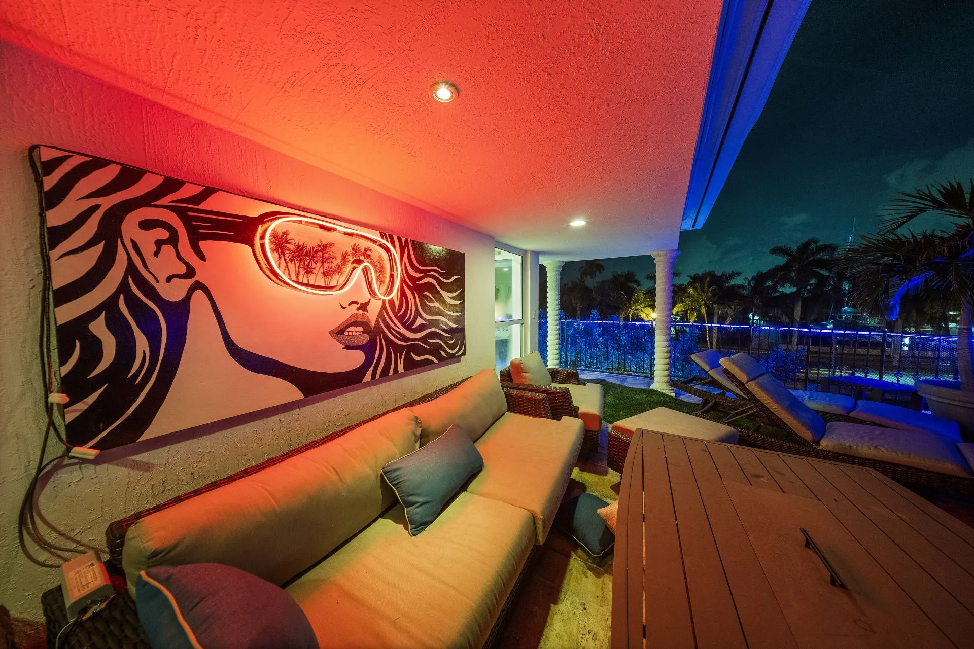 Cozy outdoor patio at night with cushioned seating, a neon-lit artwork of a woman wearing goggles reflecting palm trees, and palm trees visible beyond a railing.