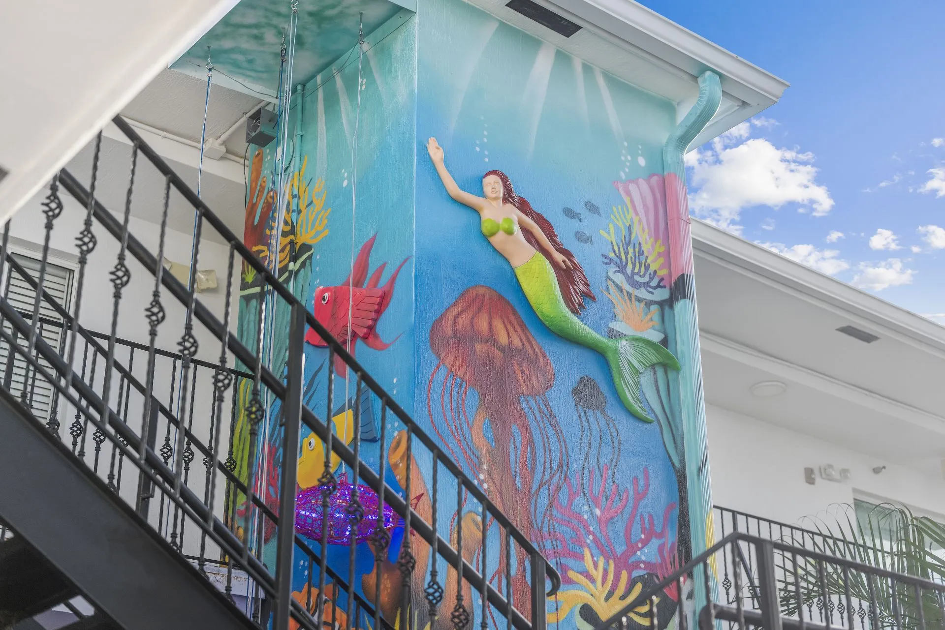 Outdoor wall mural of an underwater scene with a green-tailed mermaid, colorful fish, jellyfish, and coral near a black metal staircase.