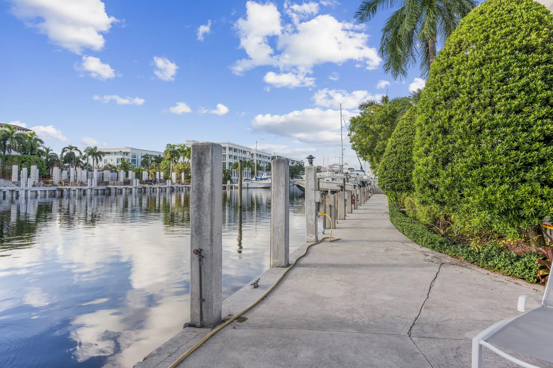 Waterfront promenade with lined concrete pillars, yachts docked in the marina, lush green bushes, and palm trees under a partly cloudy blue sky.