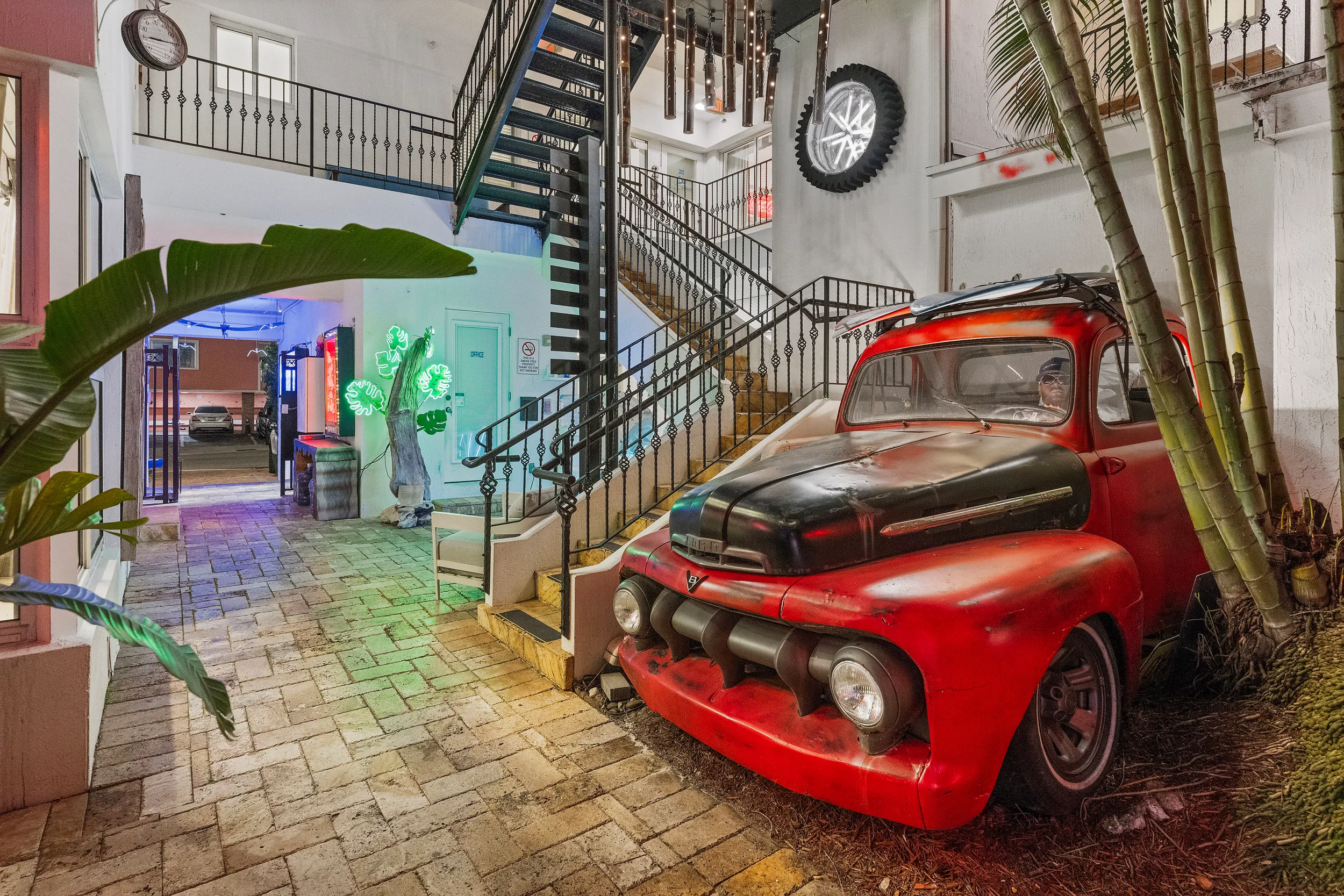 Vintage red and black truck parked indoors next to stairs with a surfboard on its roof and a mannequin driver inside.