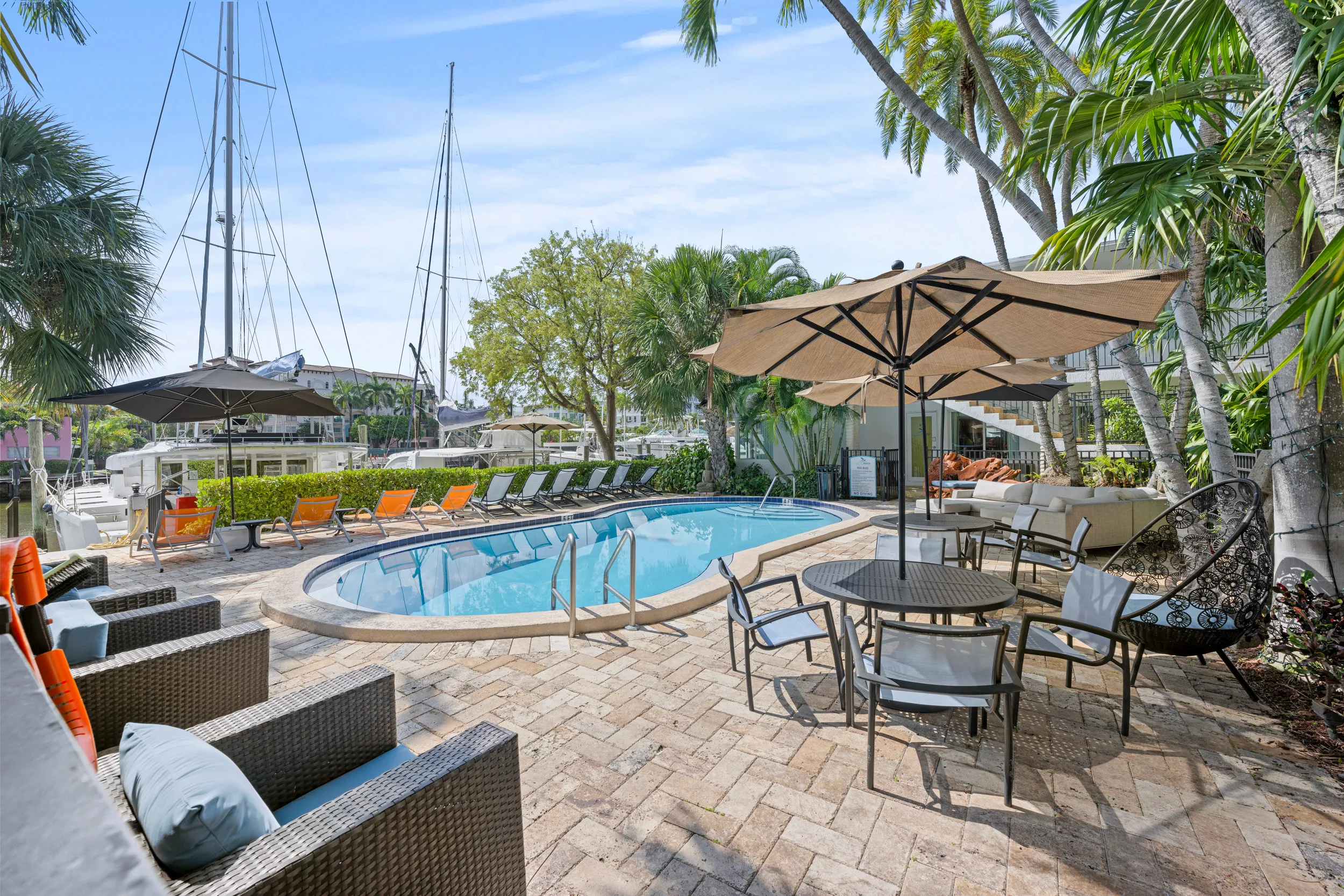 Outdoor pool area with lounge chairs, tables with umbrellas, wicker seating, palm trees, and boats docked nearby.