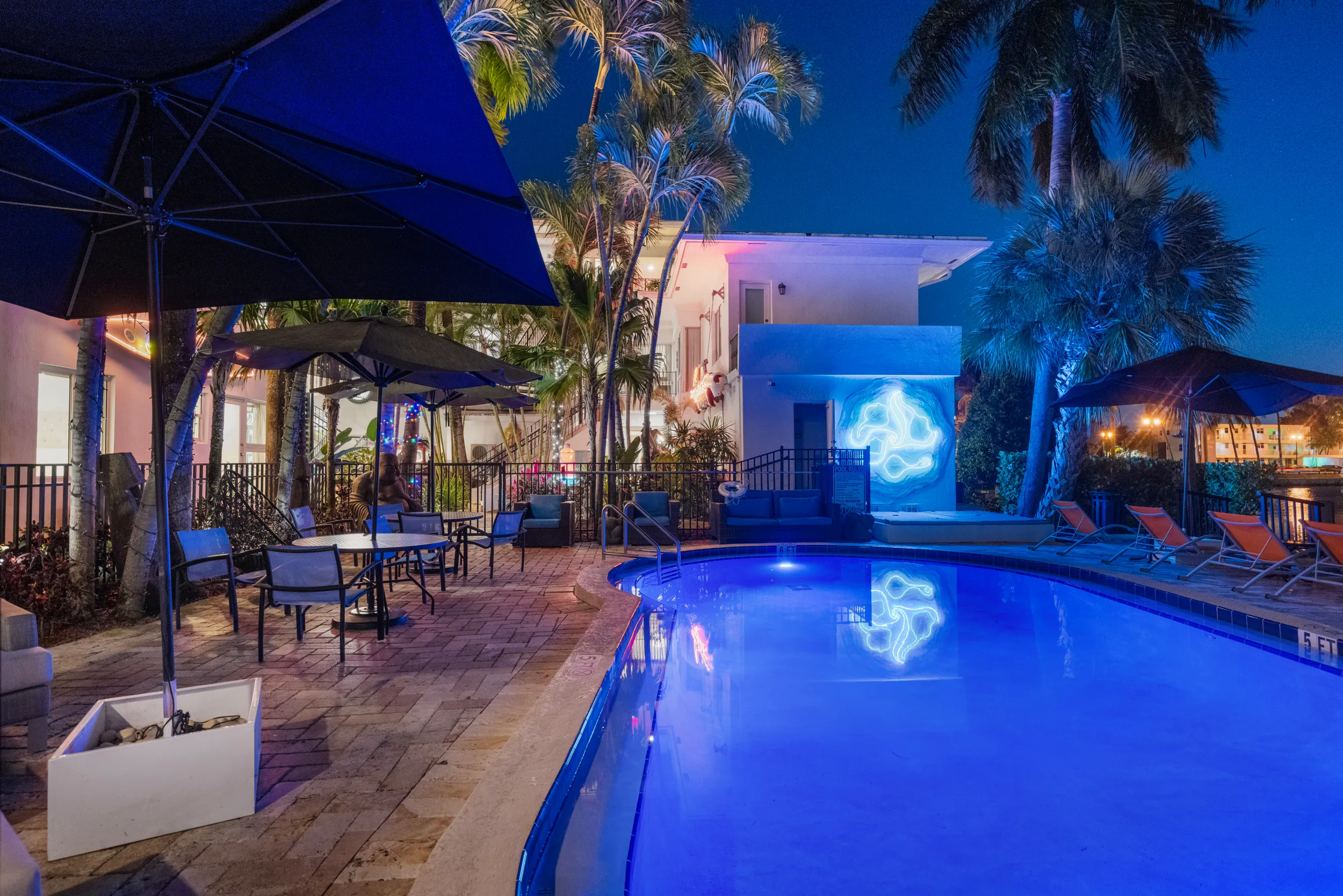 Night view of a pool area with blue lighting, lounge chairs, tables with umbrellas, palm trees, and a white building with neon wall art.