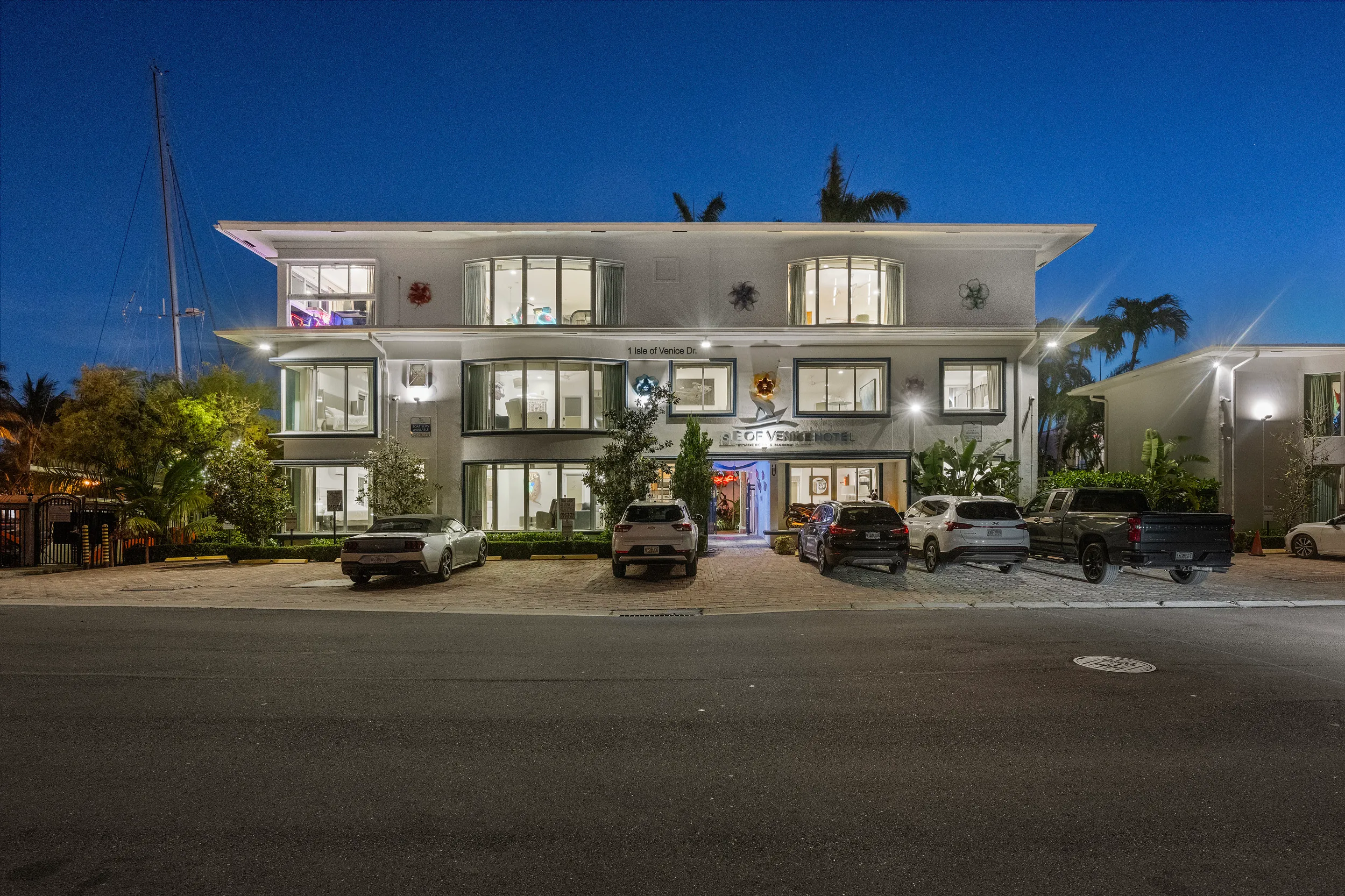 Night view of Isle of Venice Hotel with lit windows, several parked cars, and palm trees in the background.