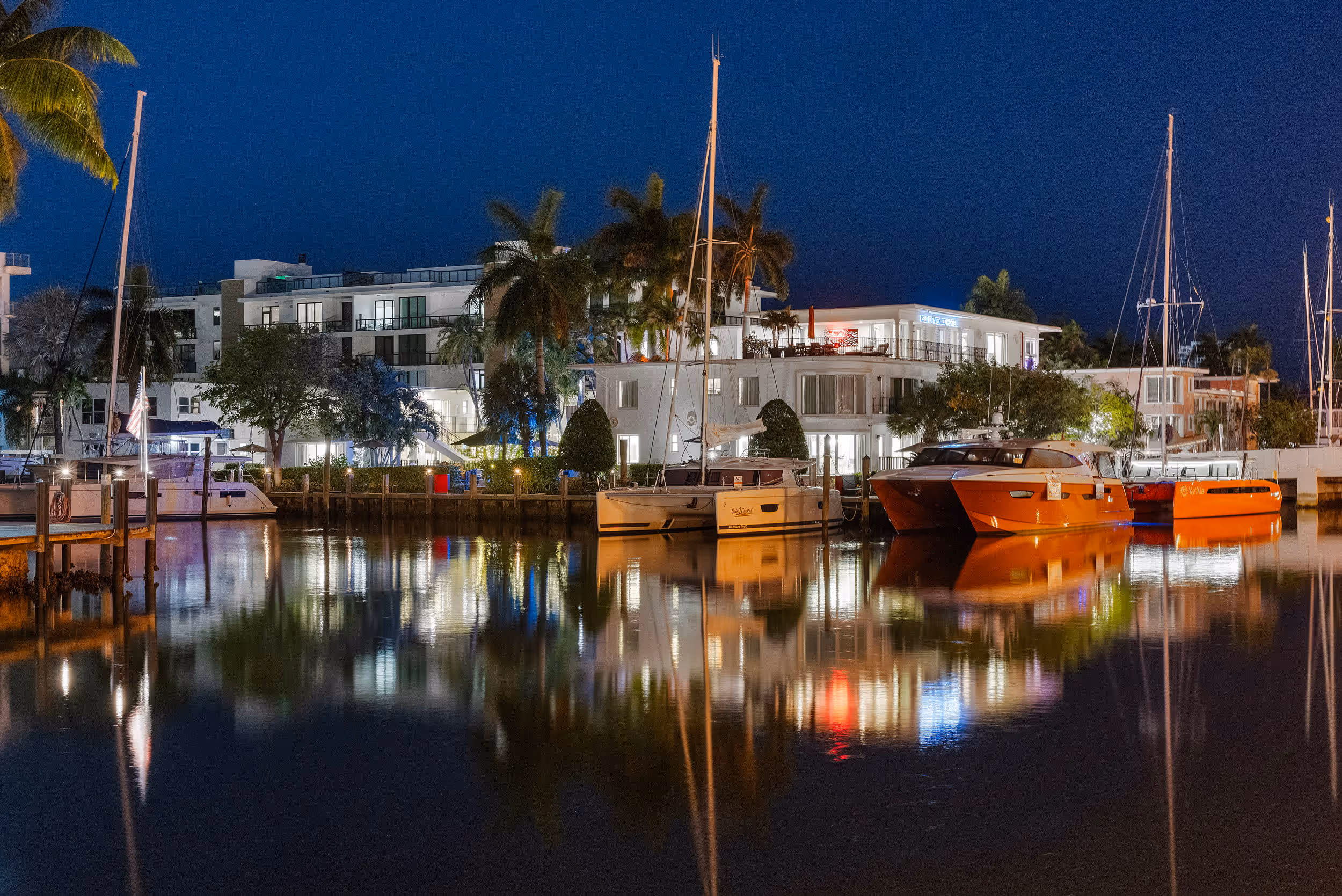 Night view of a marina with sailboats and yachts docked near palm trees and lit buildings reflecting on water.