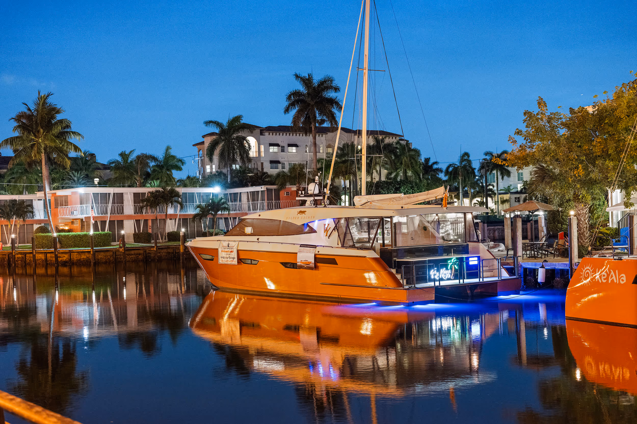 Orange yacht docked in calm marina water at dusk with palm trees and buildings in background.