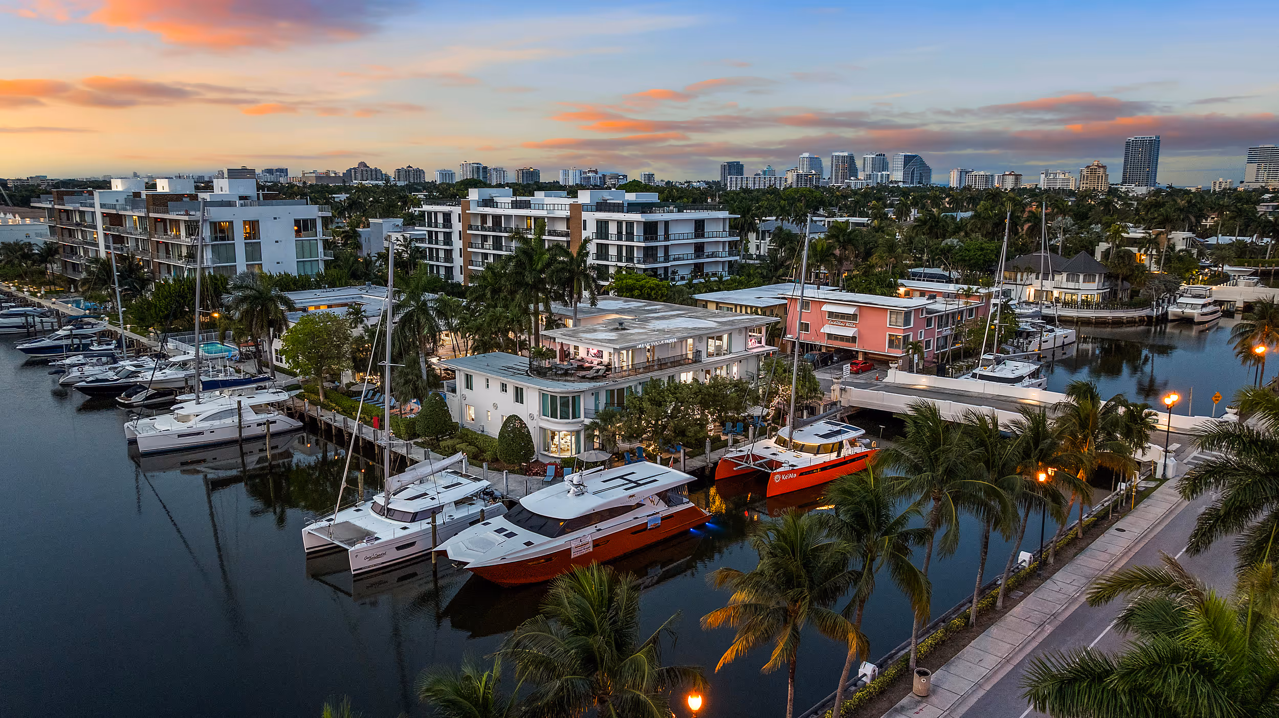 Marina at sunset with docked yachts, waterfront buildings, palm trees, and city skyline in the background.
