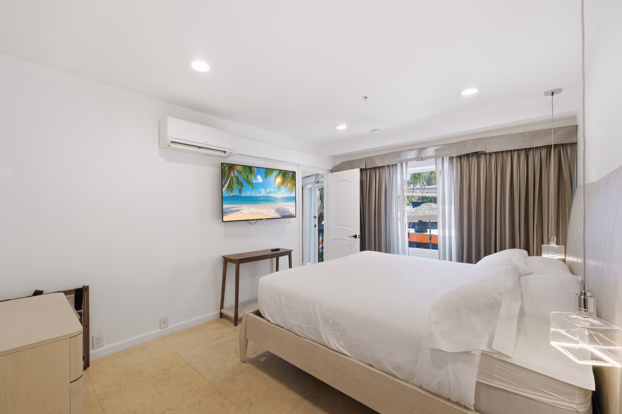Modern bedroom with white bed, mounted TV showing beach, window with curtains, and air conditioner.