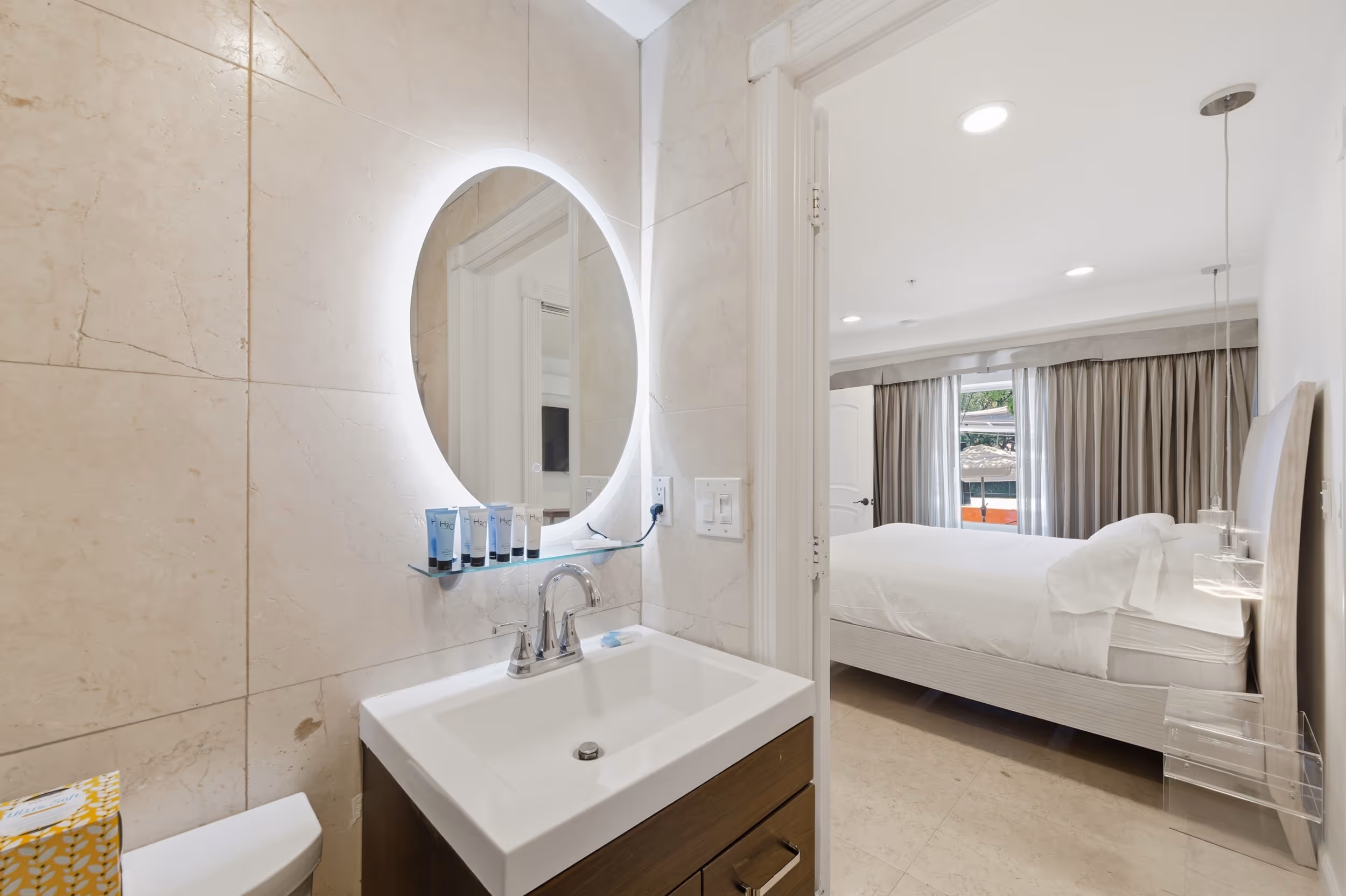 Modern bathroom sink with round backlit mirror showing a bedroom with a white bed and curtains.