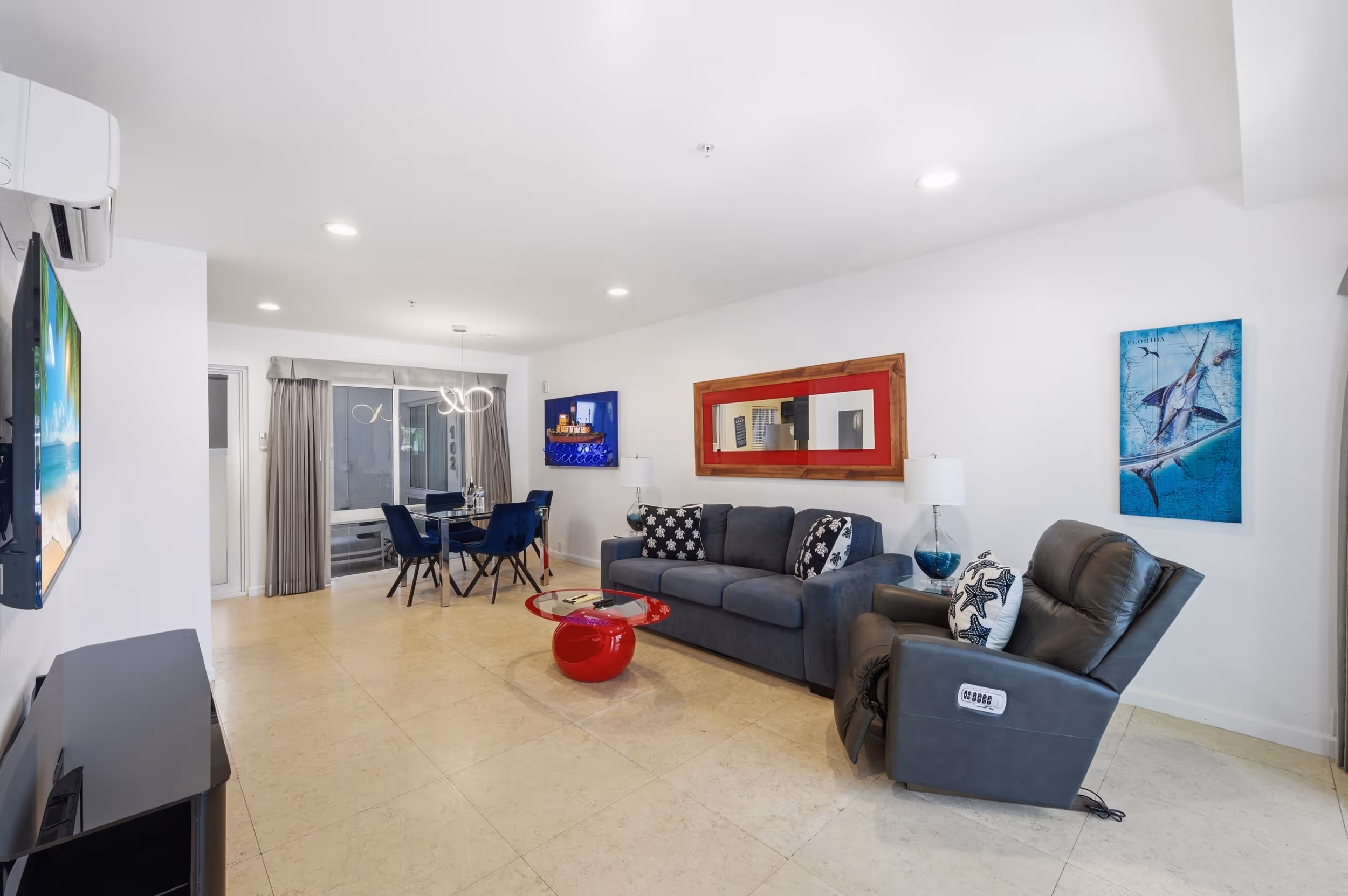 Modern living room with blue sofa, reclining chair, red glass coffee table, and dining table with blue chairs.