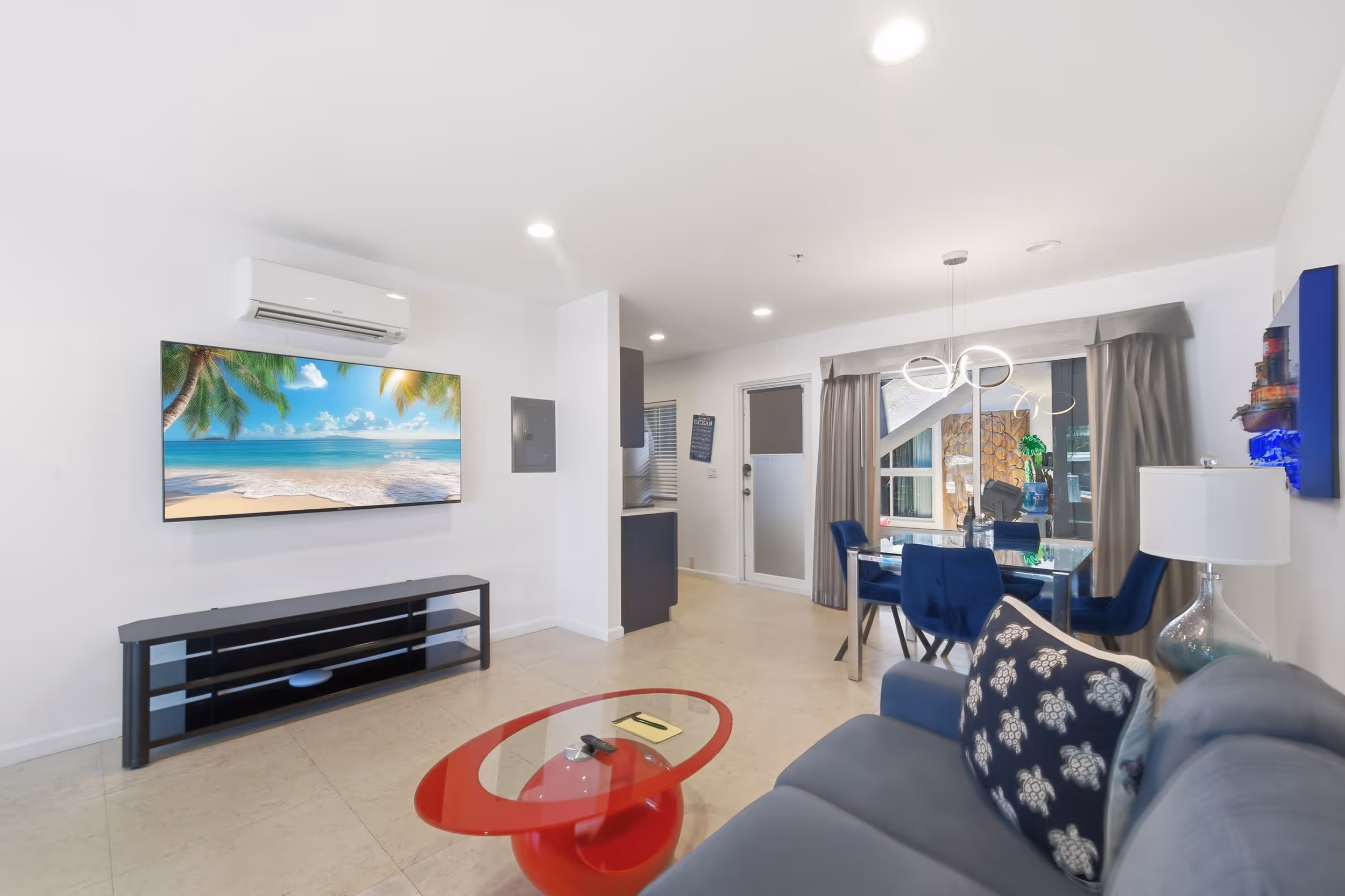 Modern living room with gray couch, red glass coffee table, TV with beach image, and blue dining chairs.