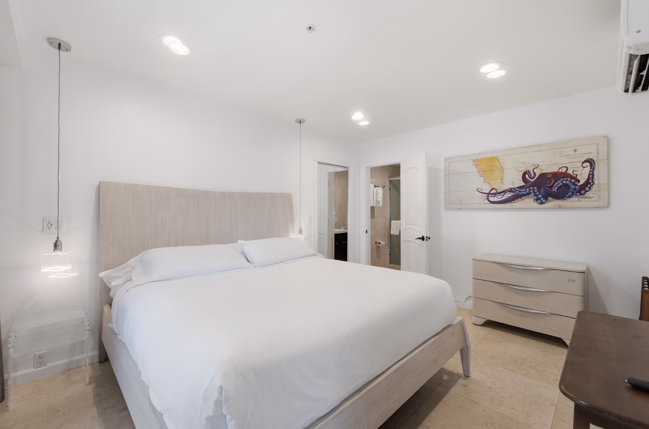 Bright bedroom with a large white bed, wooden dresser, octopus wall art, and an open door to a bathroom.