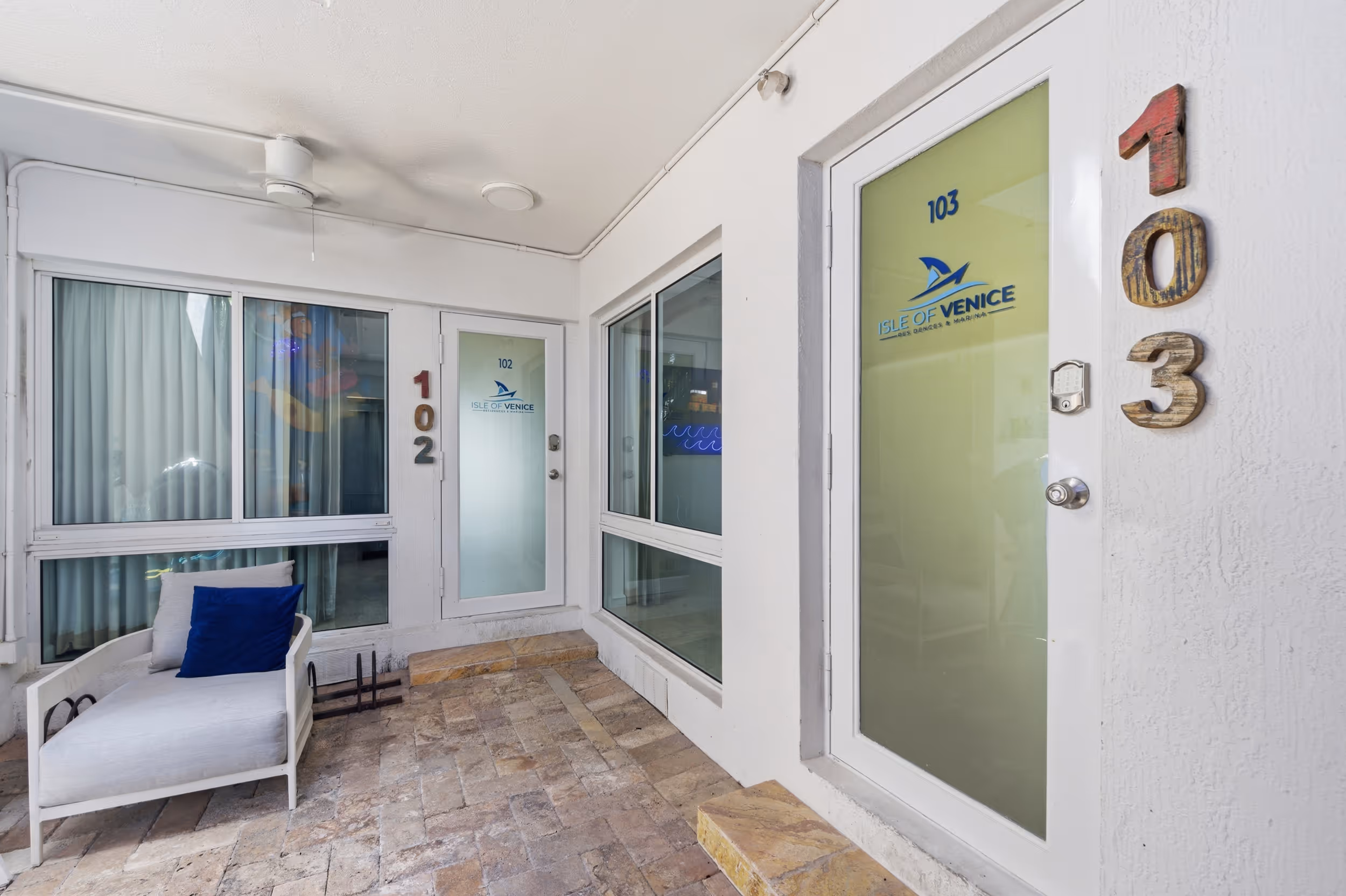 Entry doors 102 and 103 with Isle of Venice logo, white walls, and a lounge chair with blue pillow outside.