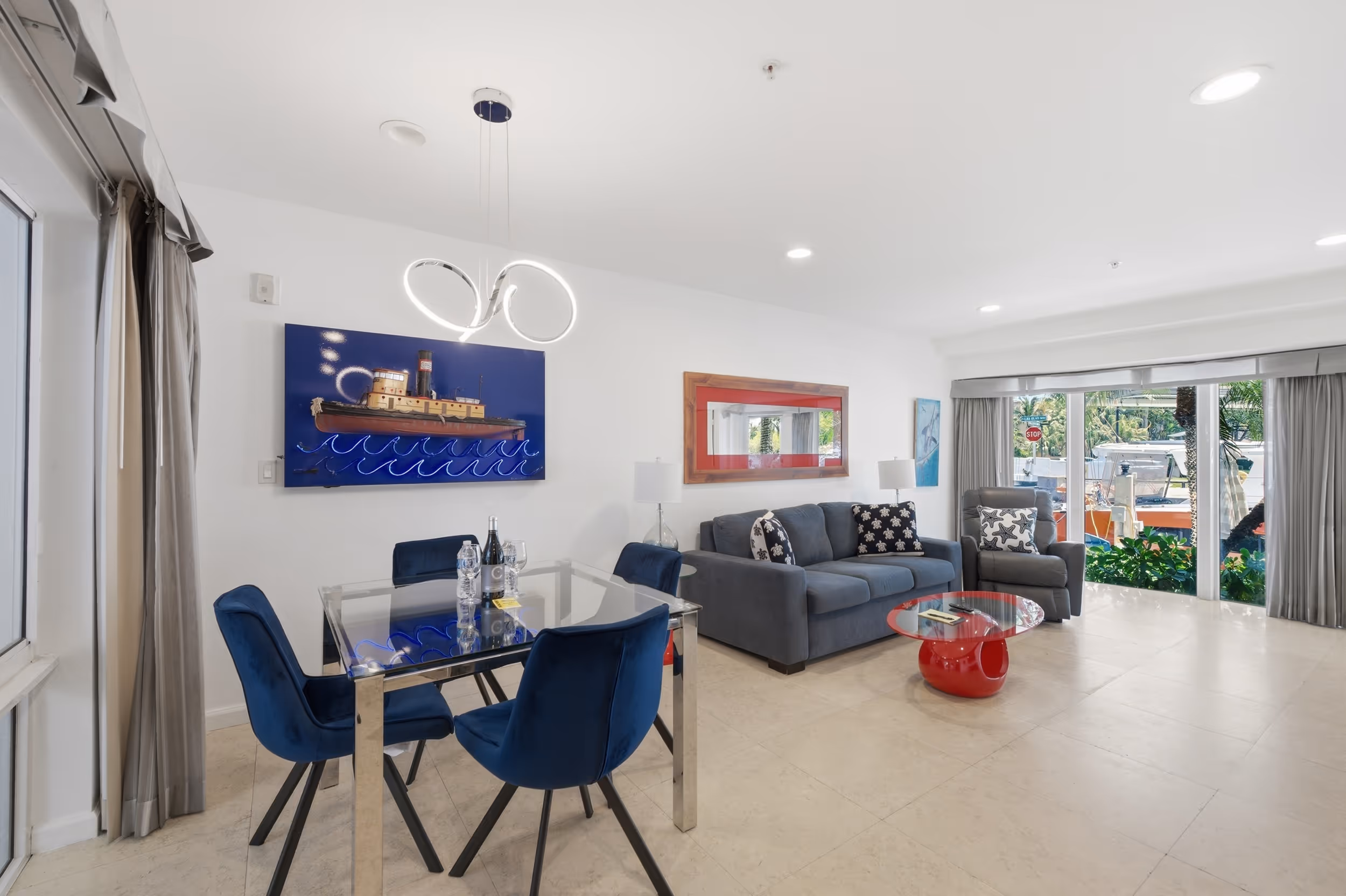 Bright living room with gray couch, blue velvet chairs, glass dining table, and large windows with outdoor view.
