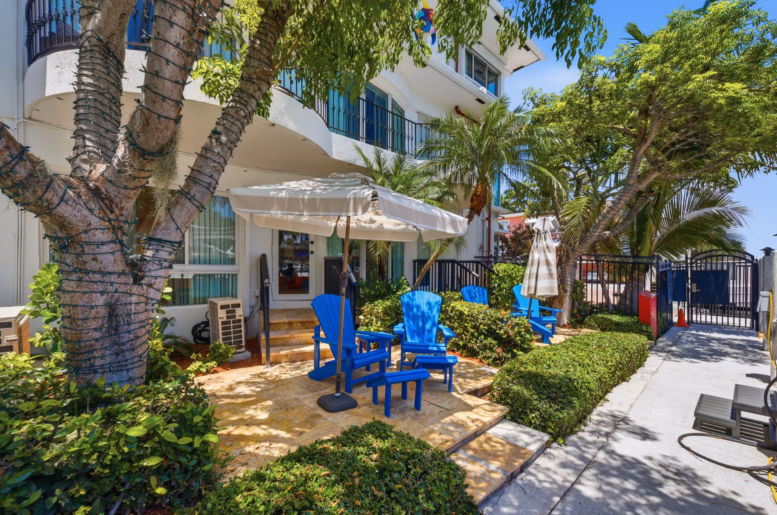 Blue Adirondack chairs under umbrellas on a patio surrounded by greenery at a white building.