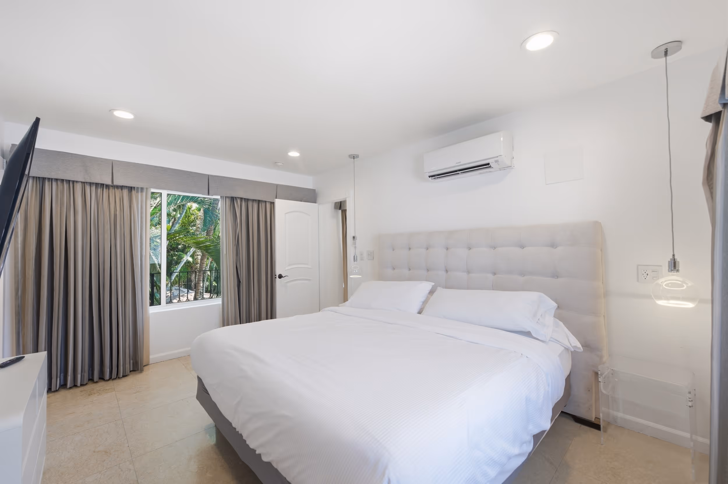 Bright bedroom with king bed, tufted headboard, pendant lights, TV, and window with curtains overlooking greenery.