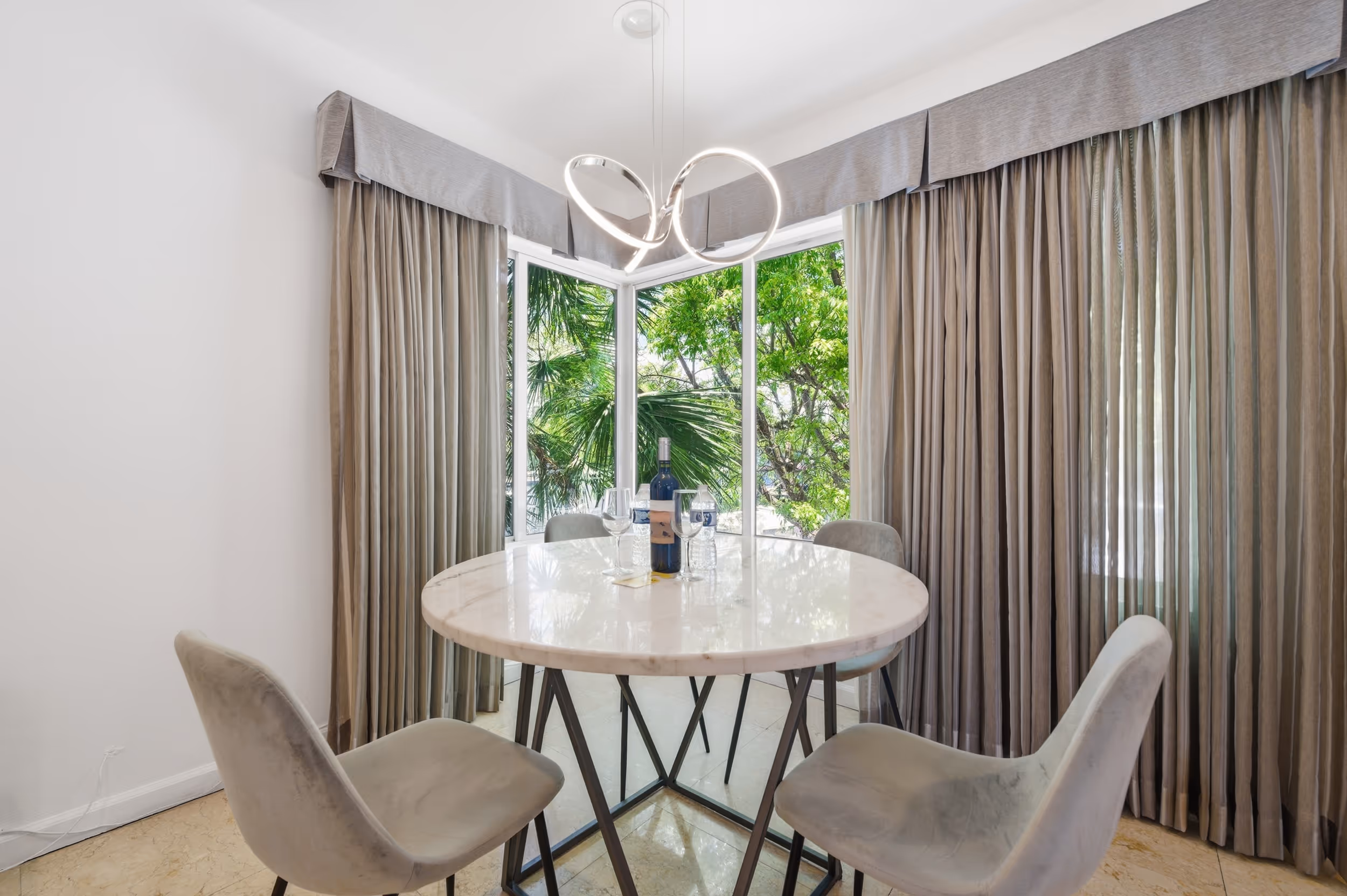 Modern dining area with round marble table, four grey chairs, and large windows with curtains.