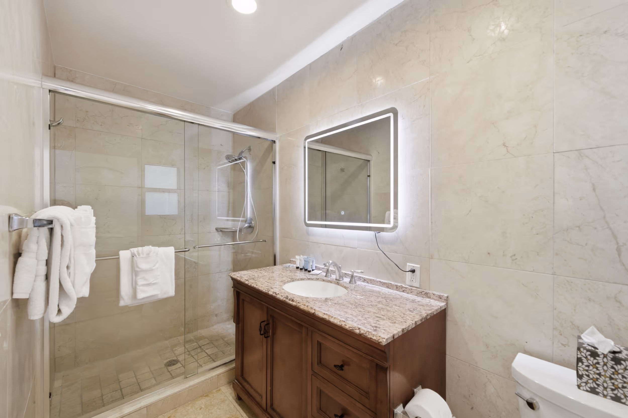 Modern bathroom with glass shower, granite countertop, wooden vanity, LED mirror, and white towels.