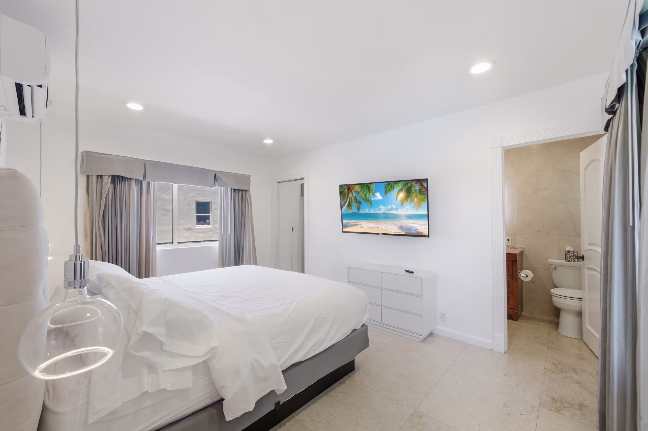 Modern white bedroom with a bed, dresser, TV showing beach scene, and open door to a bathroom.