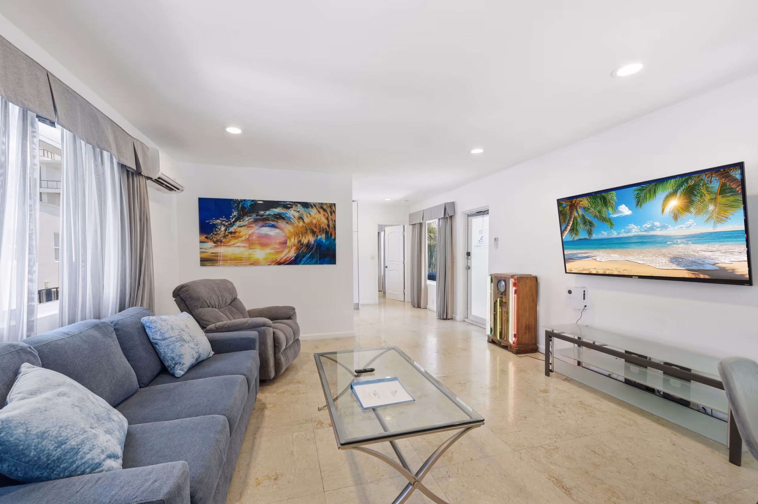 Bright living room with gray couch, recliner, glass coffee table, and a TV showing a beach scene.