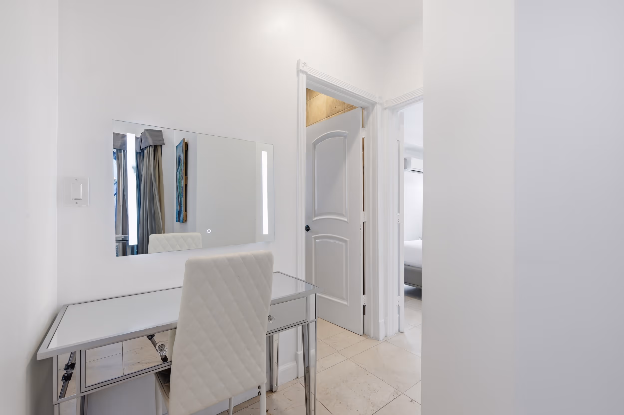 White quilted chair in front of glass desk with mirror and hallway leading to other rooms.