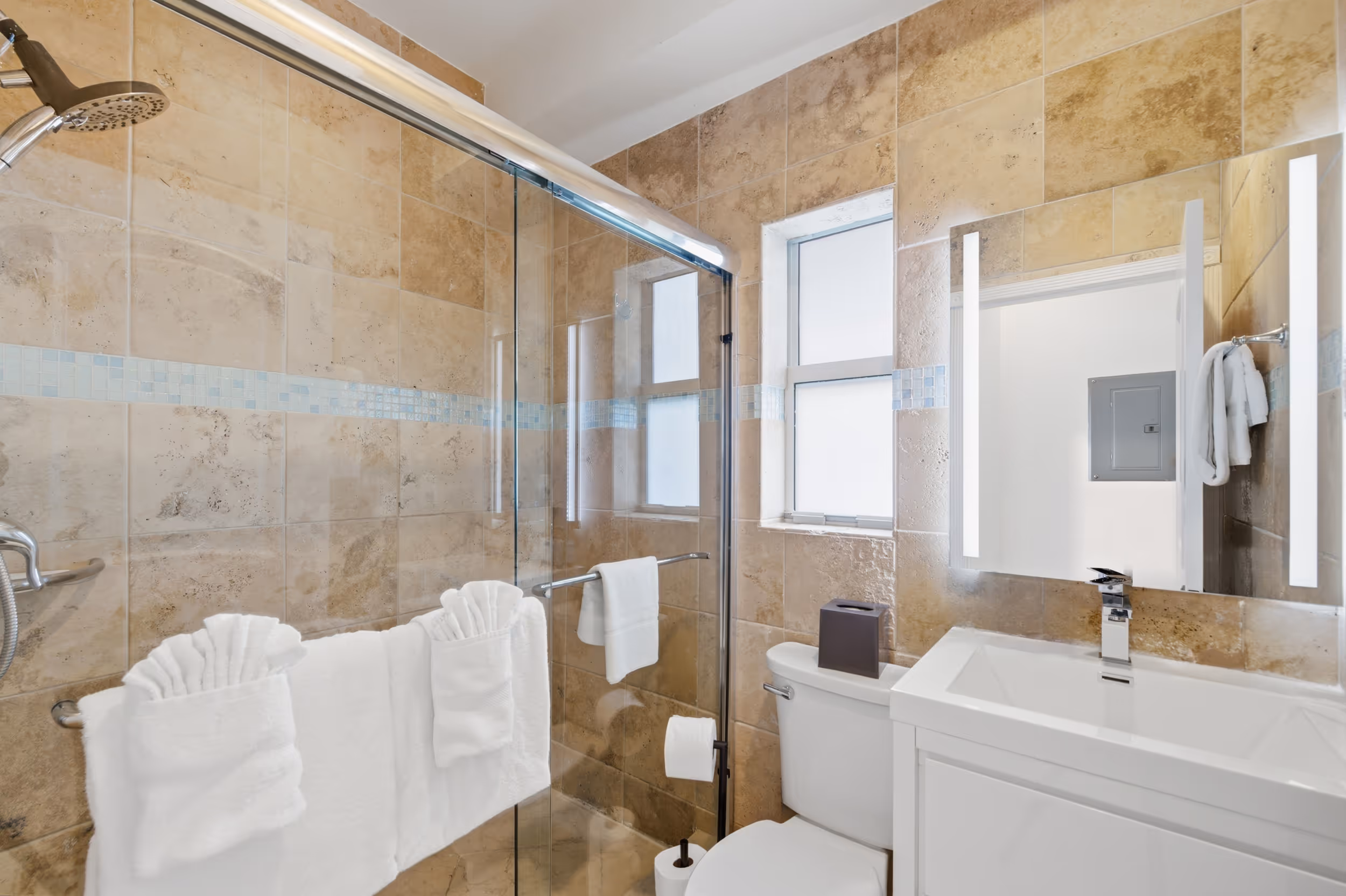 Bathroom with tan tiled walls, glass shower enclosure, white towels, toilet, and modern white sink vanity.