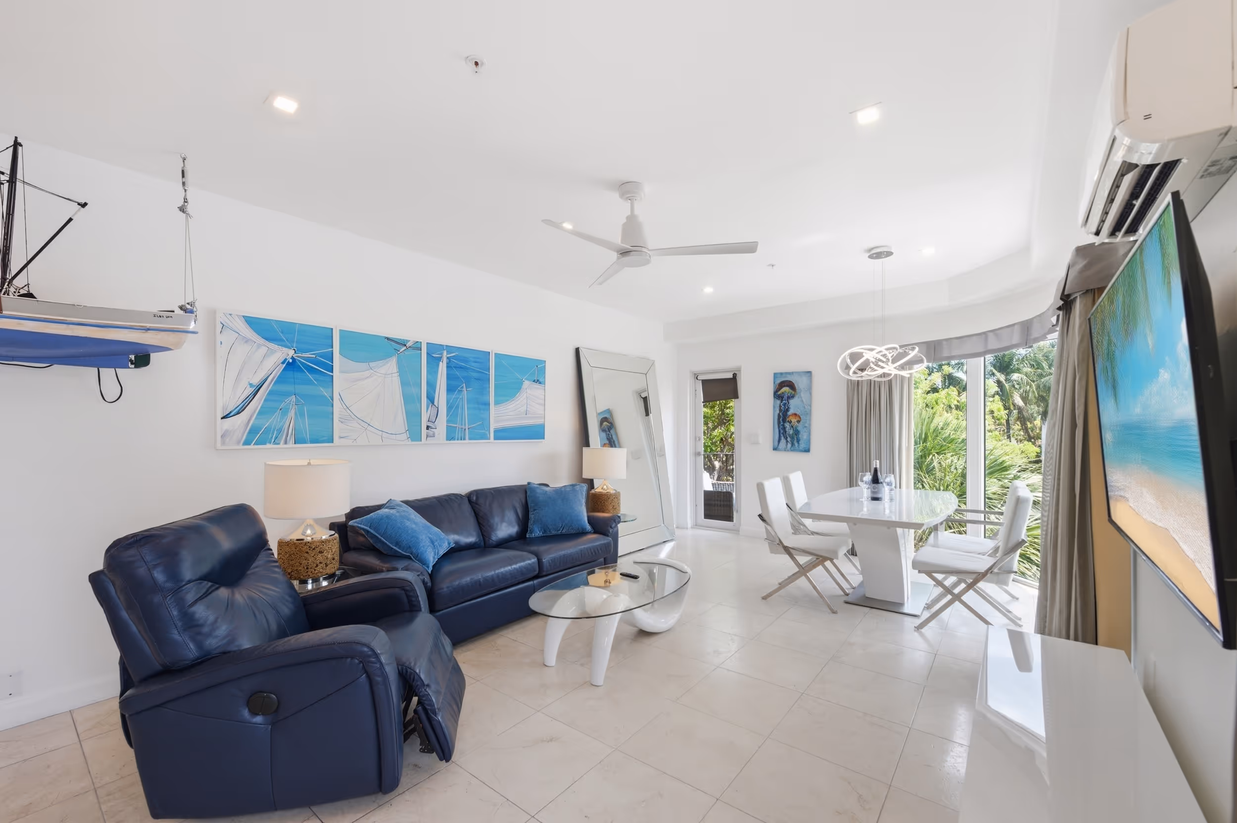 Bright living room with navy blue sofa and recliner, glass table, white dining set, and large windows.