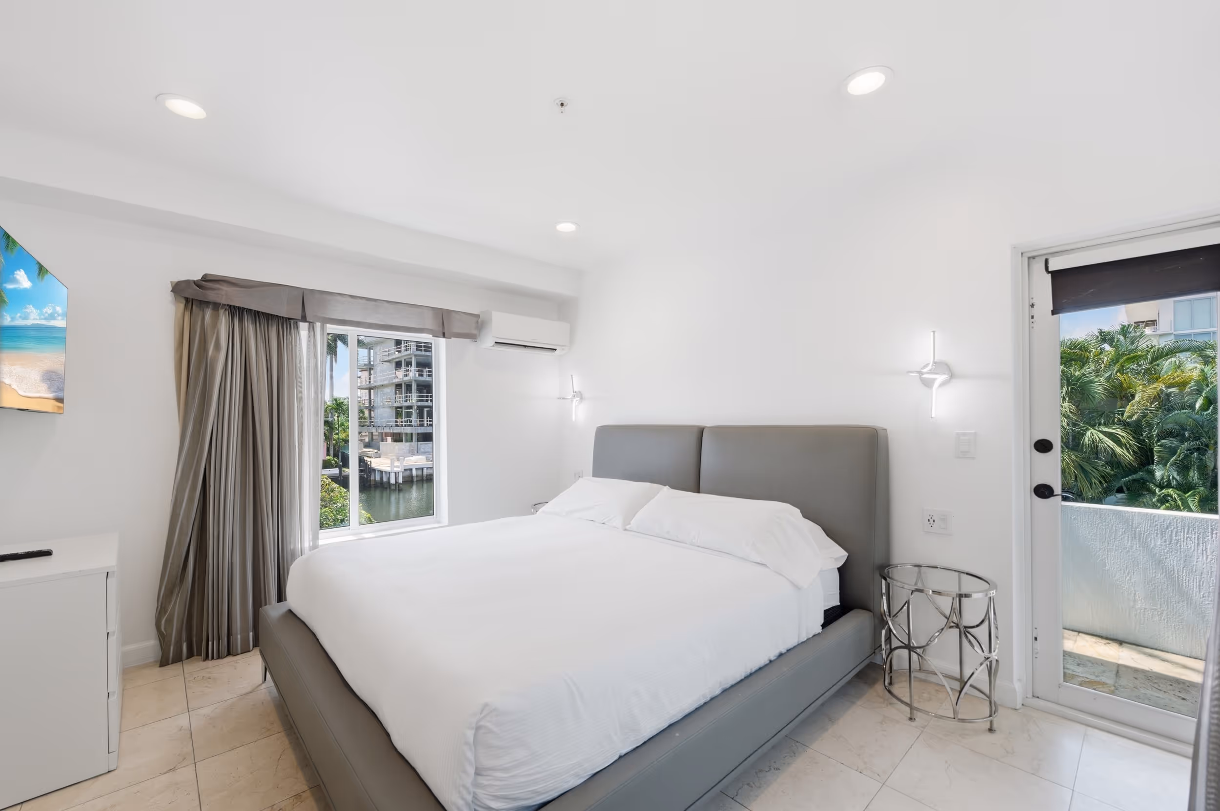 Minimalist bedroom with a large gray bed, white linens, window, balcony door, and modern lighting.