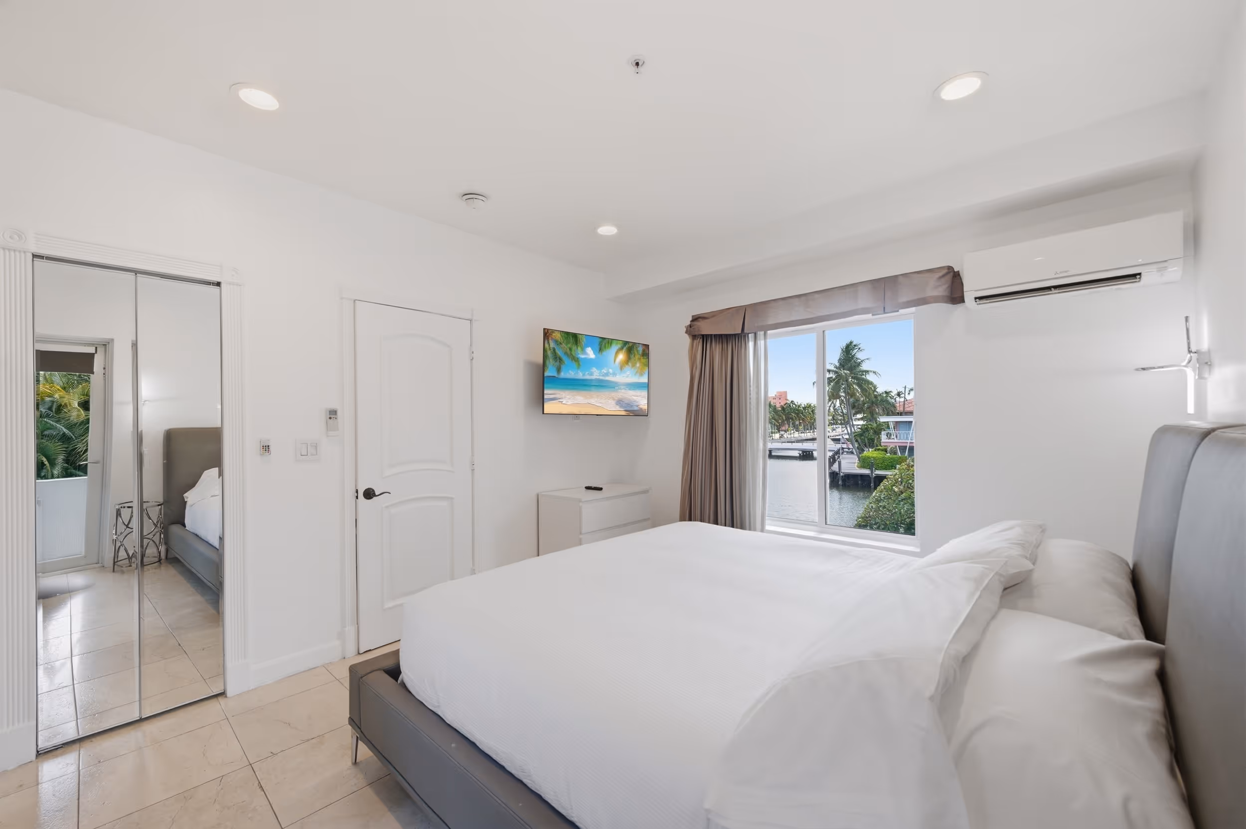 Minimalist bedroom with white linens, a wall-mounted TV, mirrored closet, and a window view of water and palm trees.