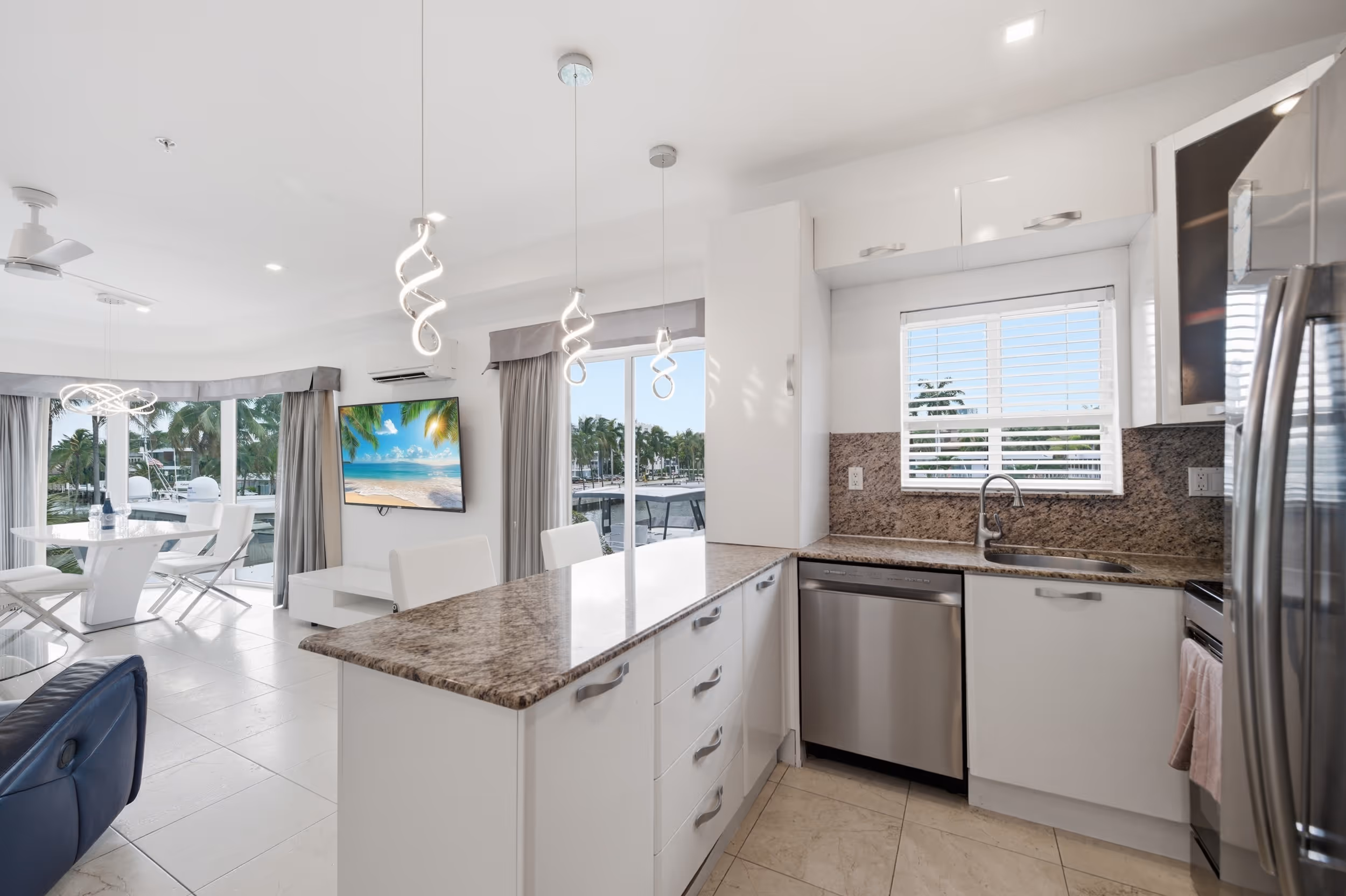 Bright modern kitchen with granite counters, stainless appliances, dining area, and large windows overlooking palm trees.