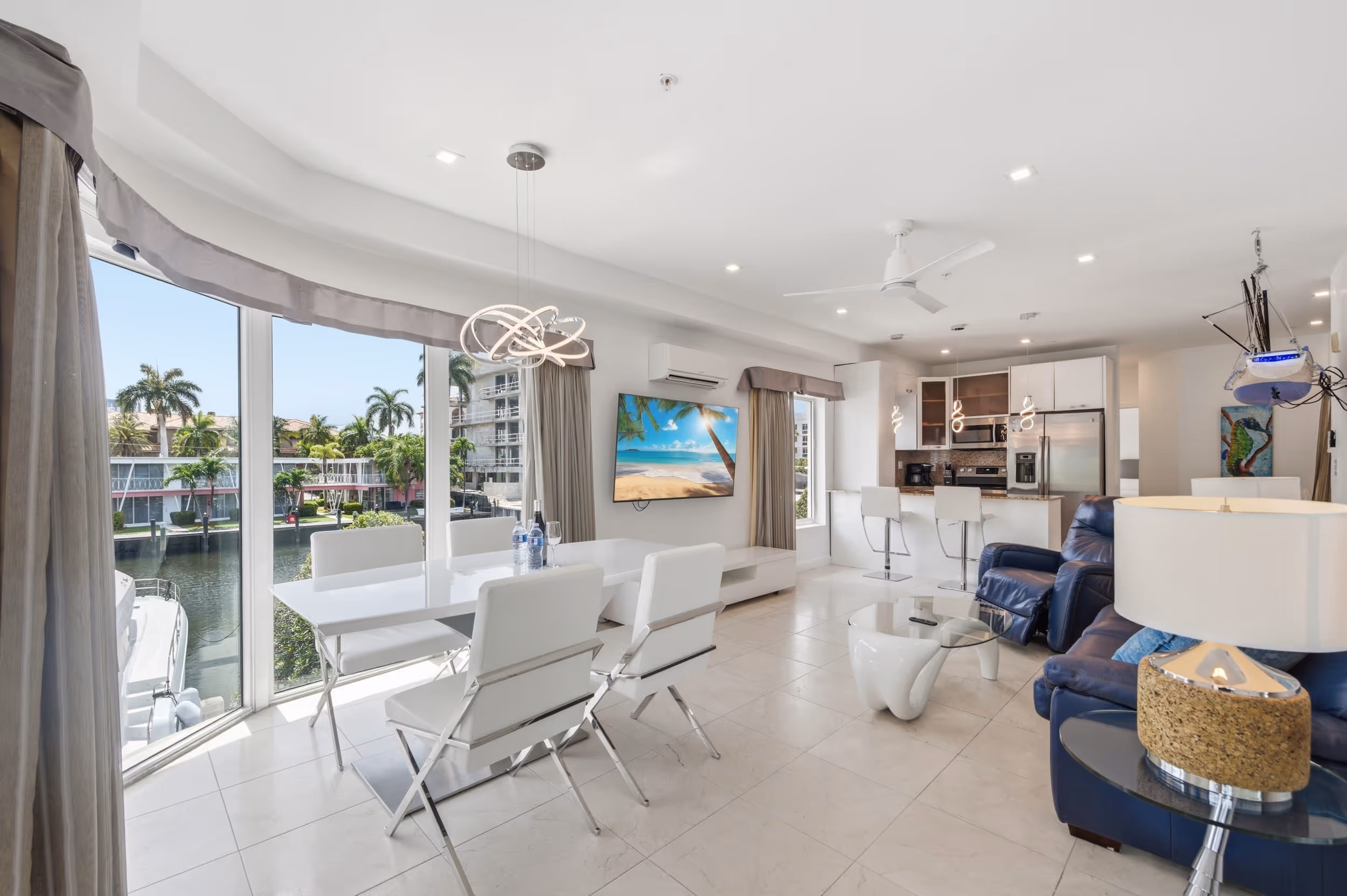 Modern living room and kitchen with white furniture, leather chairs, large windows, and canal view.