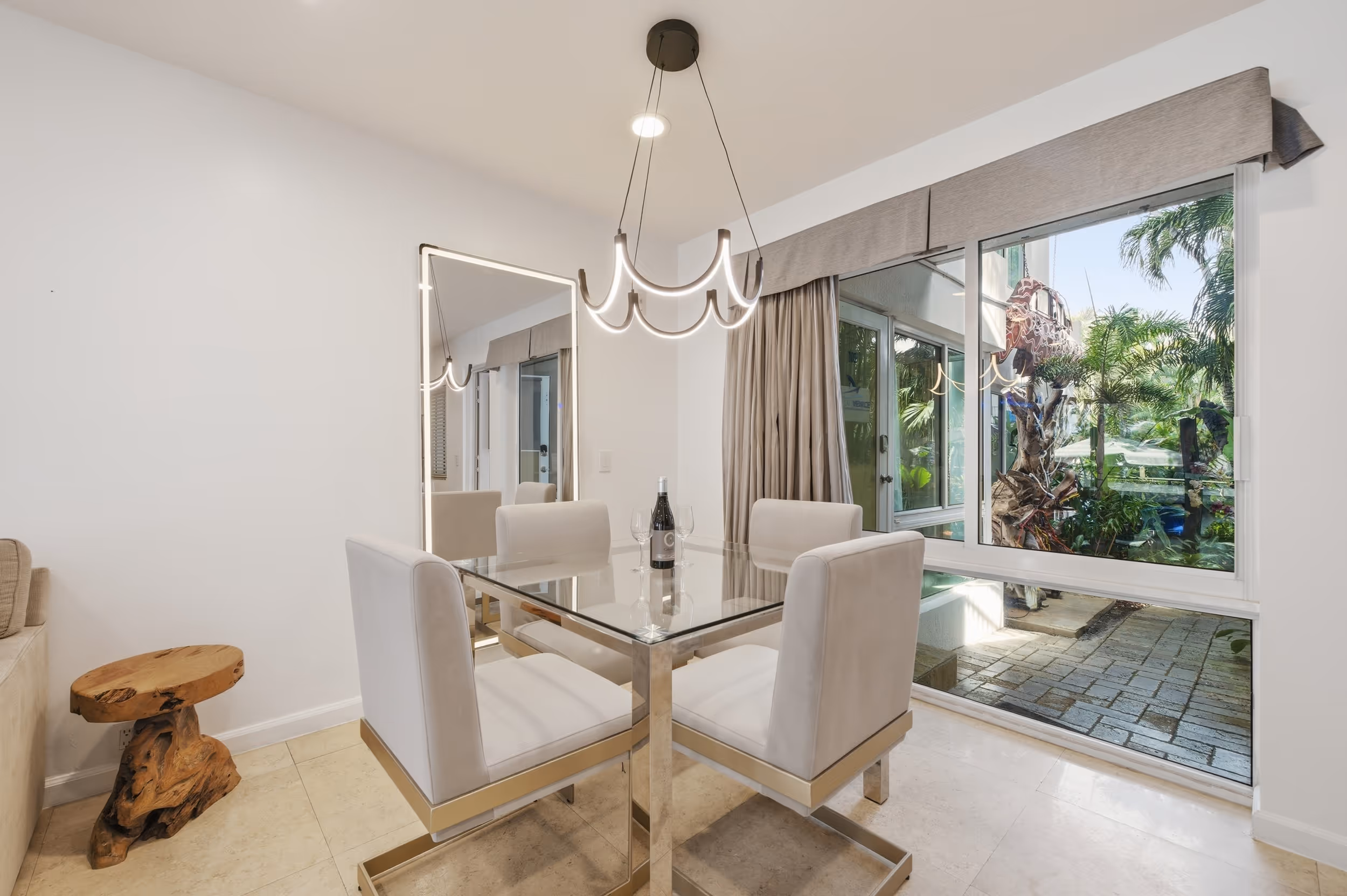 Modern dining area with glass table, four white chairs, hanging light, and large window with garden view.