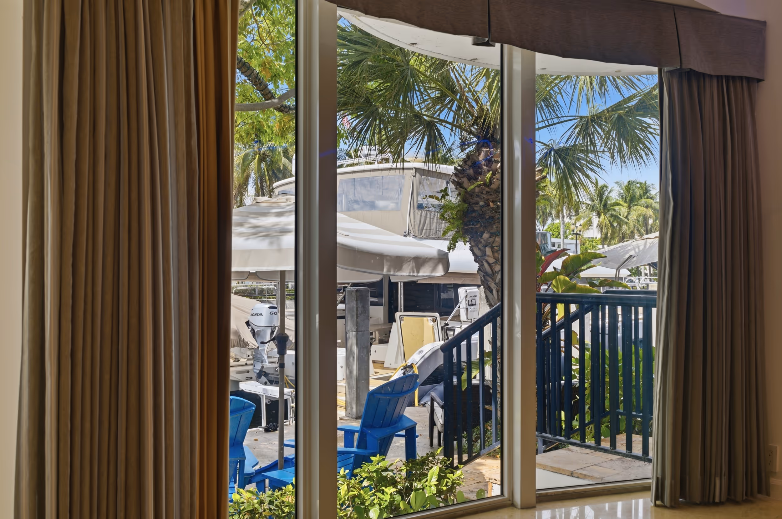 View through sliding glass door of a dock with blue chairs, palm trees, and a boat outside.