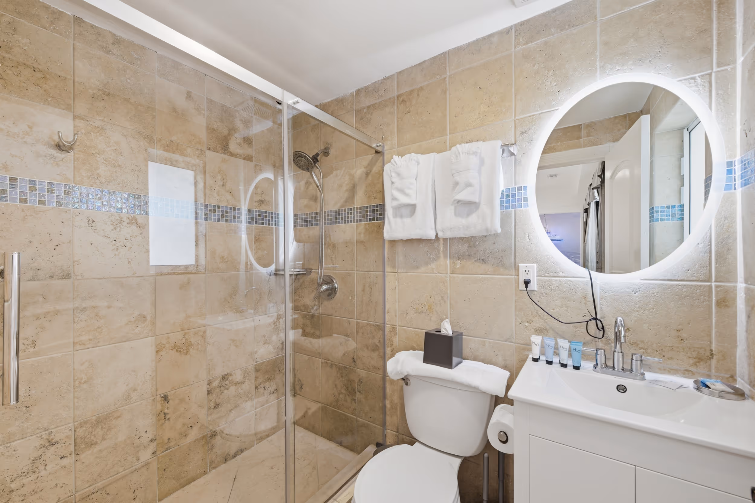 Bathroom with beige tiled walls, glass shower, white toilet, vanity with illuminated round mirror.