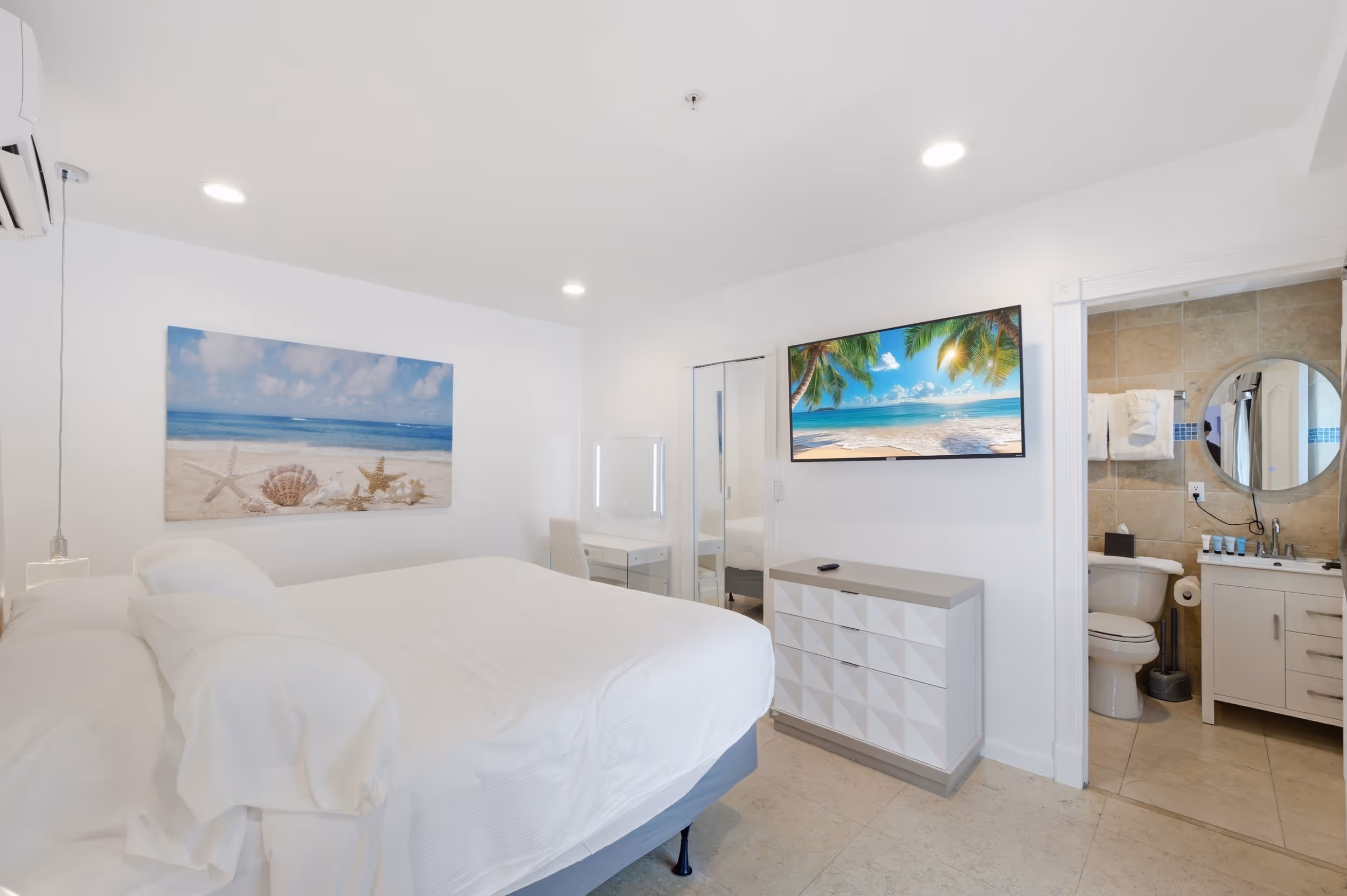 Bright modern hotel room with white bedding, beach artwork, TV, dresser, and bathroom with tiled walls.