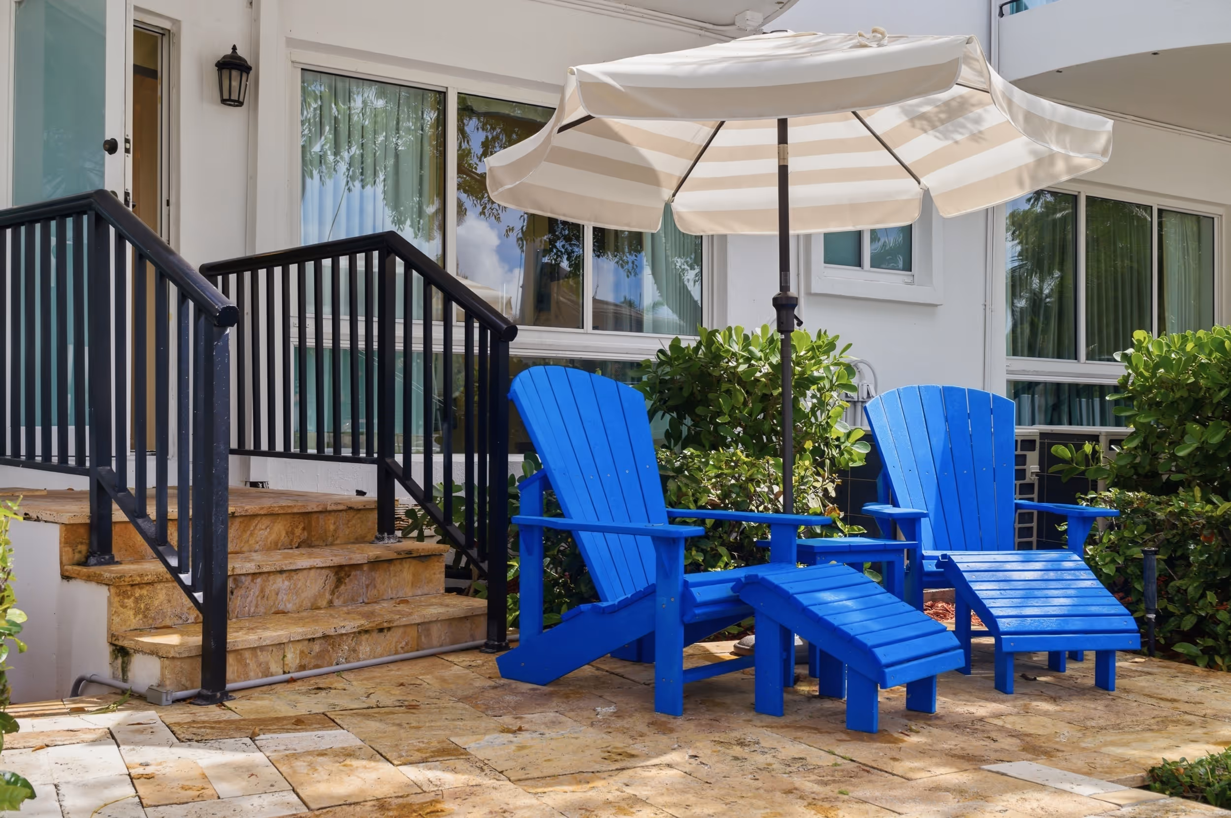 Two blue lounge chairs with footrests under a large white-striped patio umbrella by a house porch.