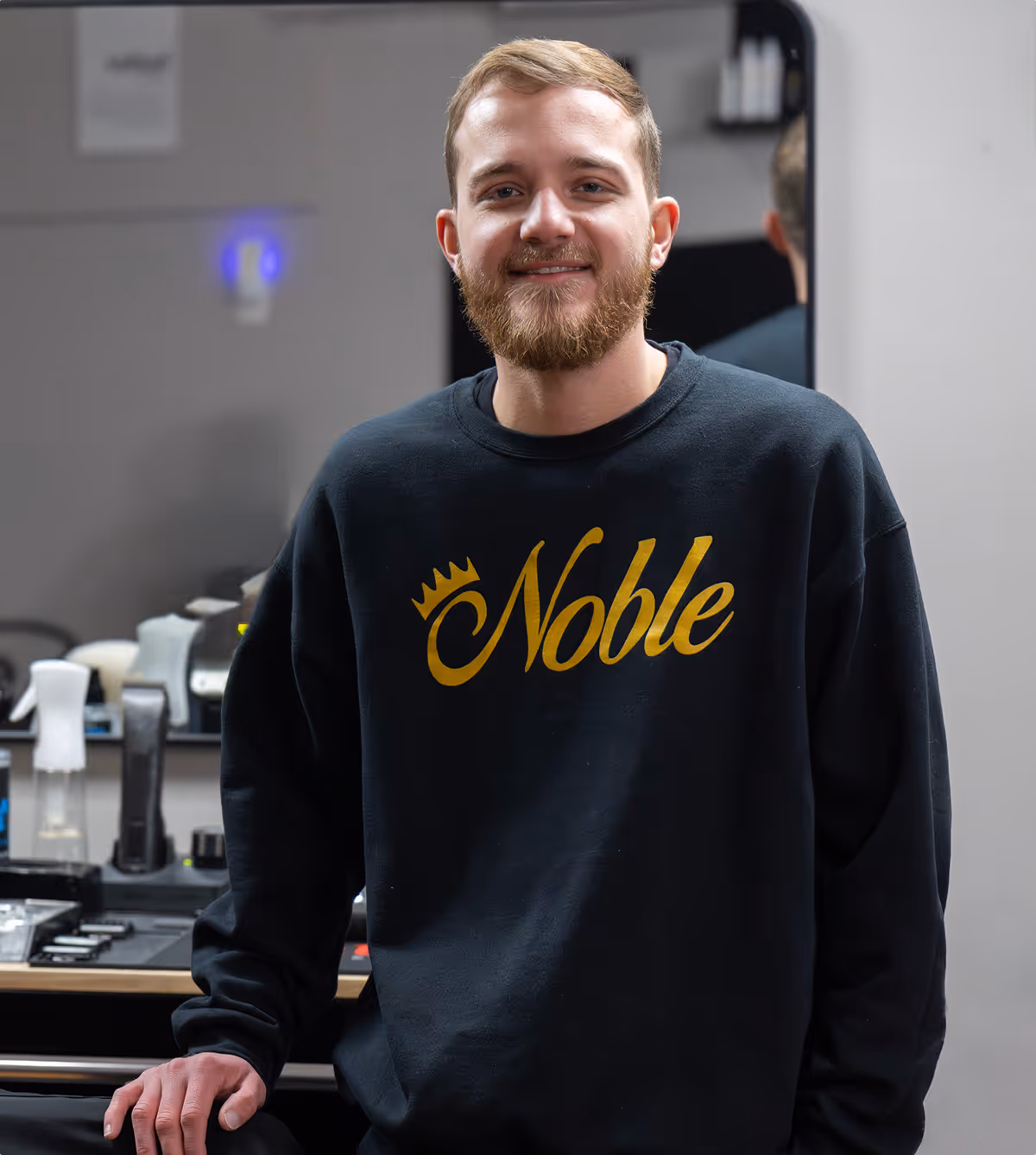 Bearded man in a black Noble sweatshirt standing in a barbershop with grooming tools behind him.