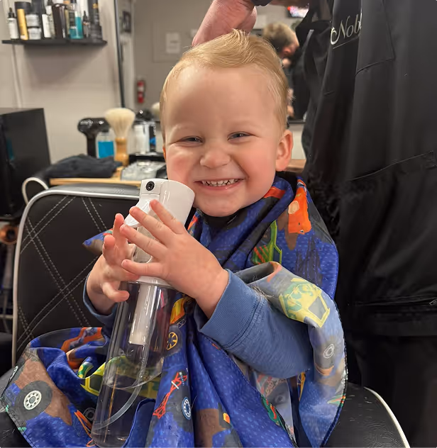 Smiling toddler sitting in salon chair wearing haircut cape and holding spray bottle.