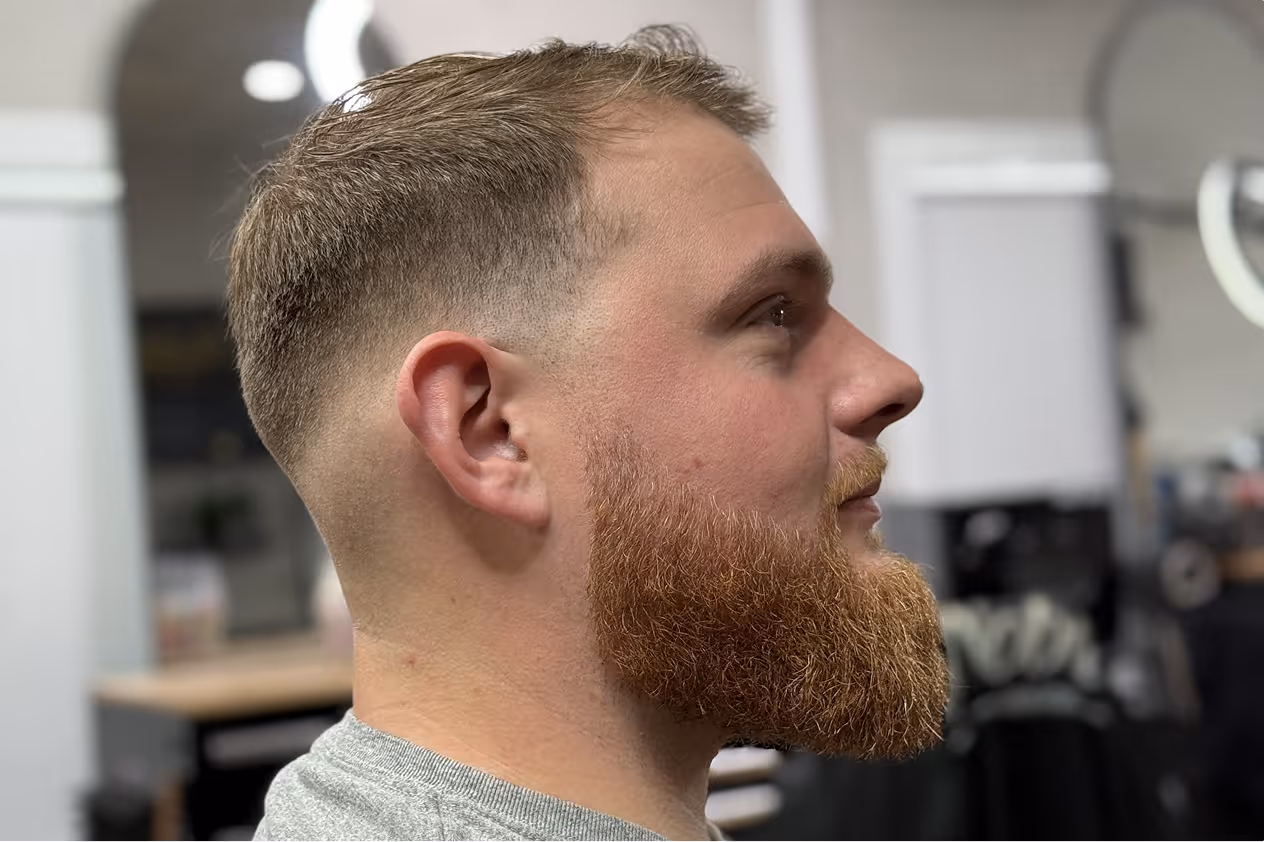 Side profile of a man with a tapered fade haircut and a full trimmed reddish beard.