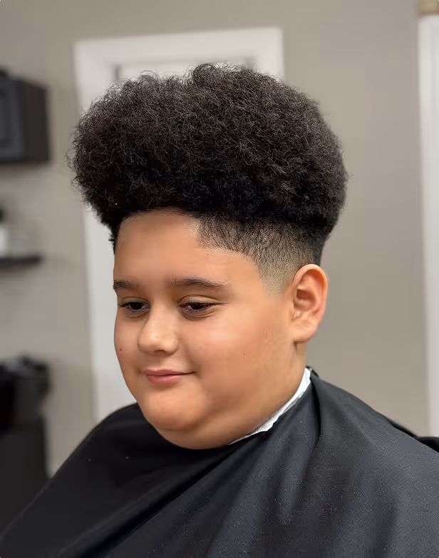 Young boy with a high-top afro hairstyle and a fade haircut wearing a black barber cape.