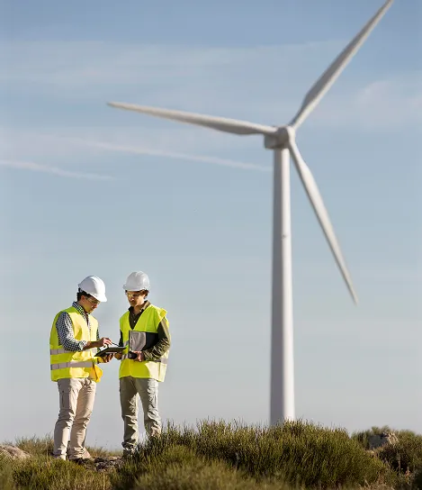Two men in yellow vests standing next to a wind turbine.