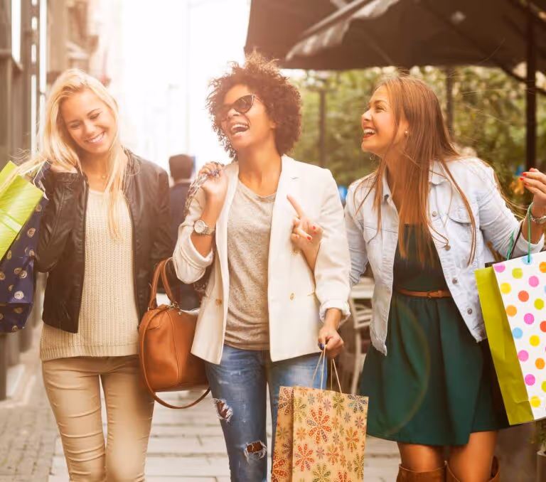 Three women smiling, carrying shopping bags, walking outdoors on a city street in daylight.