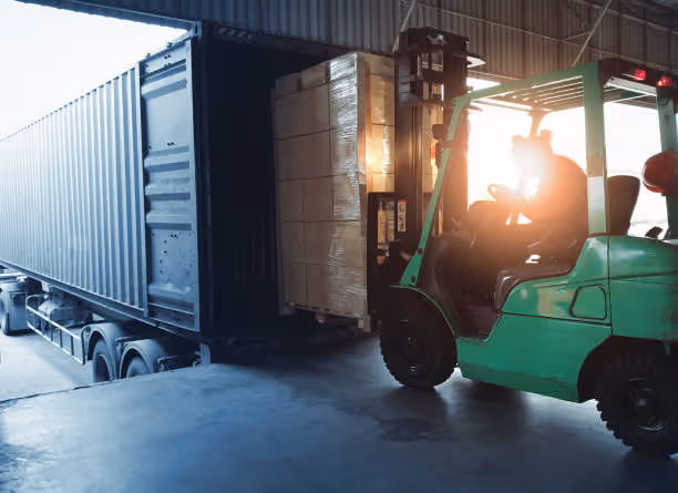 Forklift loading palletized boxes into a shipping container inside a warehouse with sunlight in background.
