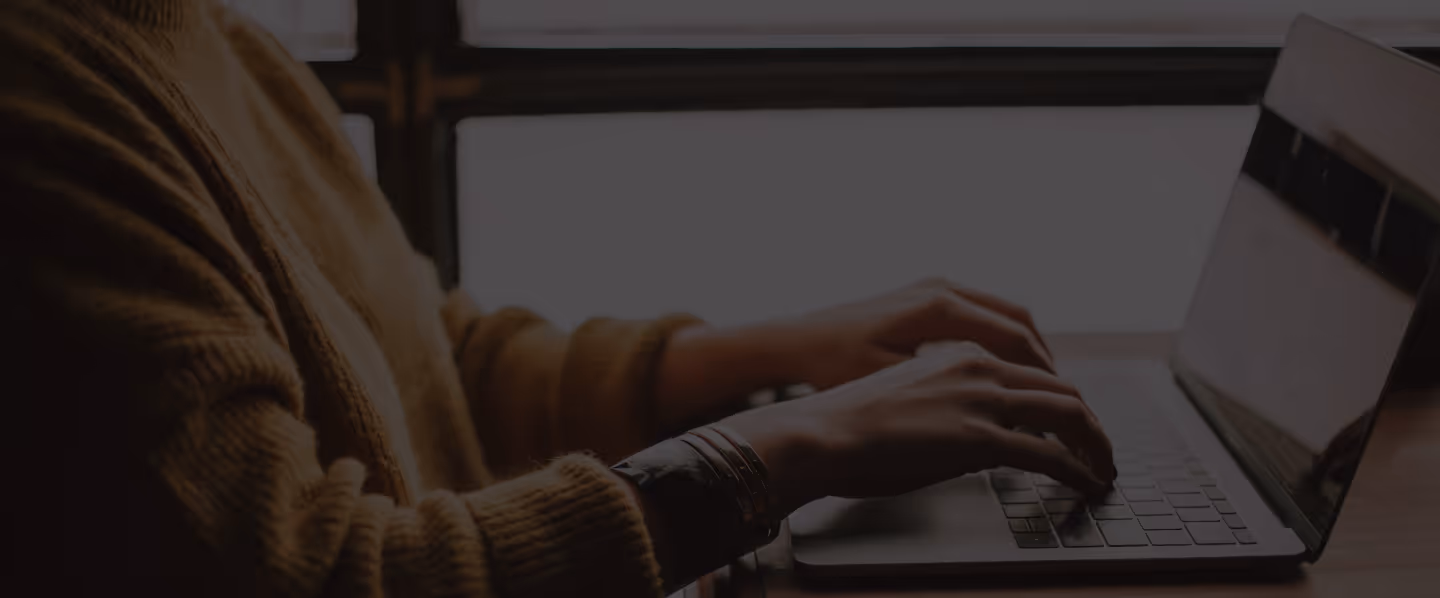 Person in brown sweater typing on a laptop keyboard by a wooden table near a window.