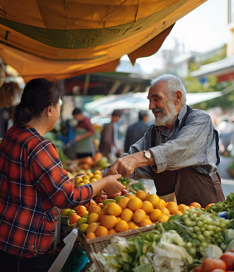 Elderly male vendor handing citrus fruits to a female customer at an outdoor market stall.