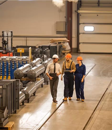Three workers with hard hats walk and discuss inside a spacious industrial factory with machinery.