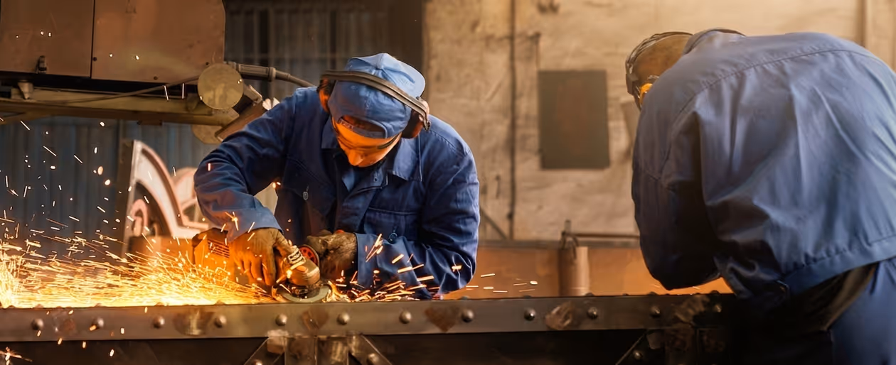 Two factory workers in blue uniforms grinding metal causing bright sparks in industrial workshop.